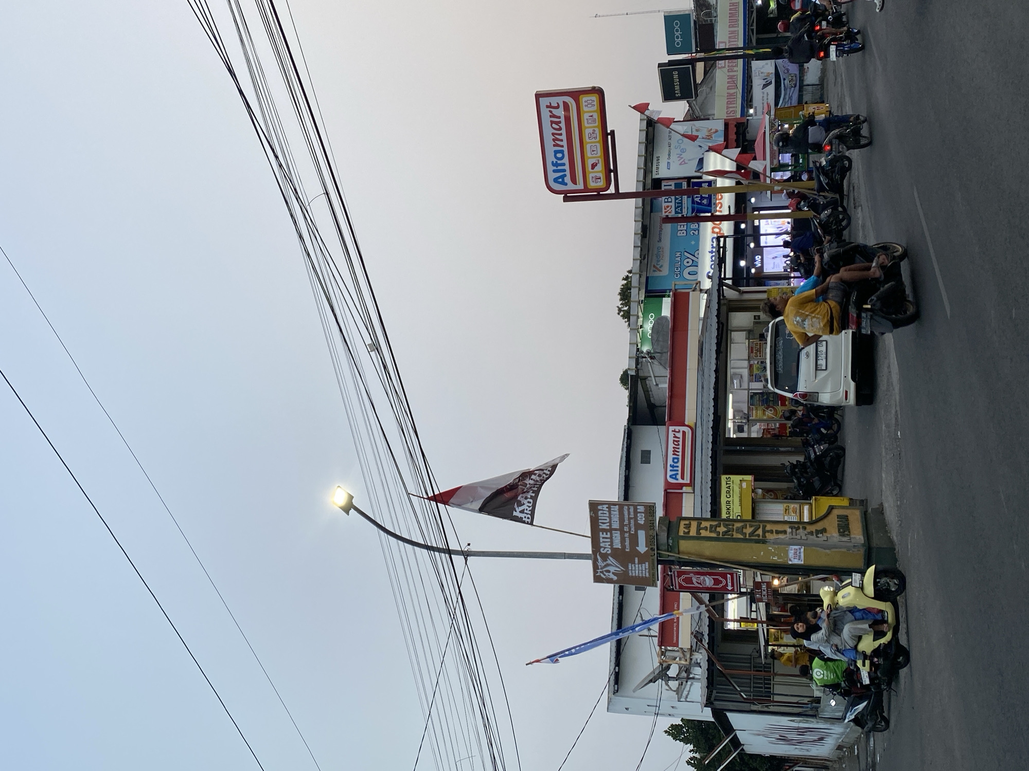 The image captures a street scene in Kasihan, Indonesia, characterized by a warm, overcast sky and a moderate amount of traffic. In the foreground, several motorcycles are visible, with one carrying a woman and child in a light yellow scooter. A man in a yellow t-shirt is riding another motorcycle behind them. Further down the street, more motorcycles are parked or in motion.
Dominating the mid-ground is an Alfamart convenience store, easily identifiable by its prominent red and white signage. In front of the store, a tall pole holds several flags, including a red and white Indonesian flag and another flag with text that is partially obscured. Below the flags, a sign advertises "SATE KUDA" (Horse Satay) with directions indicating a location in Kasihan. Adjacent to the Alfamart, other businesses are visible with various signage, including advertisements for mobile phone brands like Samsung and Oppo, and a red banner that reads "CICILAN 0%".
The environment appears to be an urban or suburban area with buildings lining the street and electrical wires overhead. The lighting suggests it might be late afternoon or early evening. The overall impression is a typical street view of a commercial area in Indonesia.
