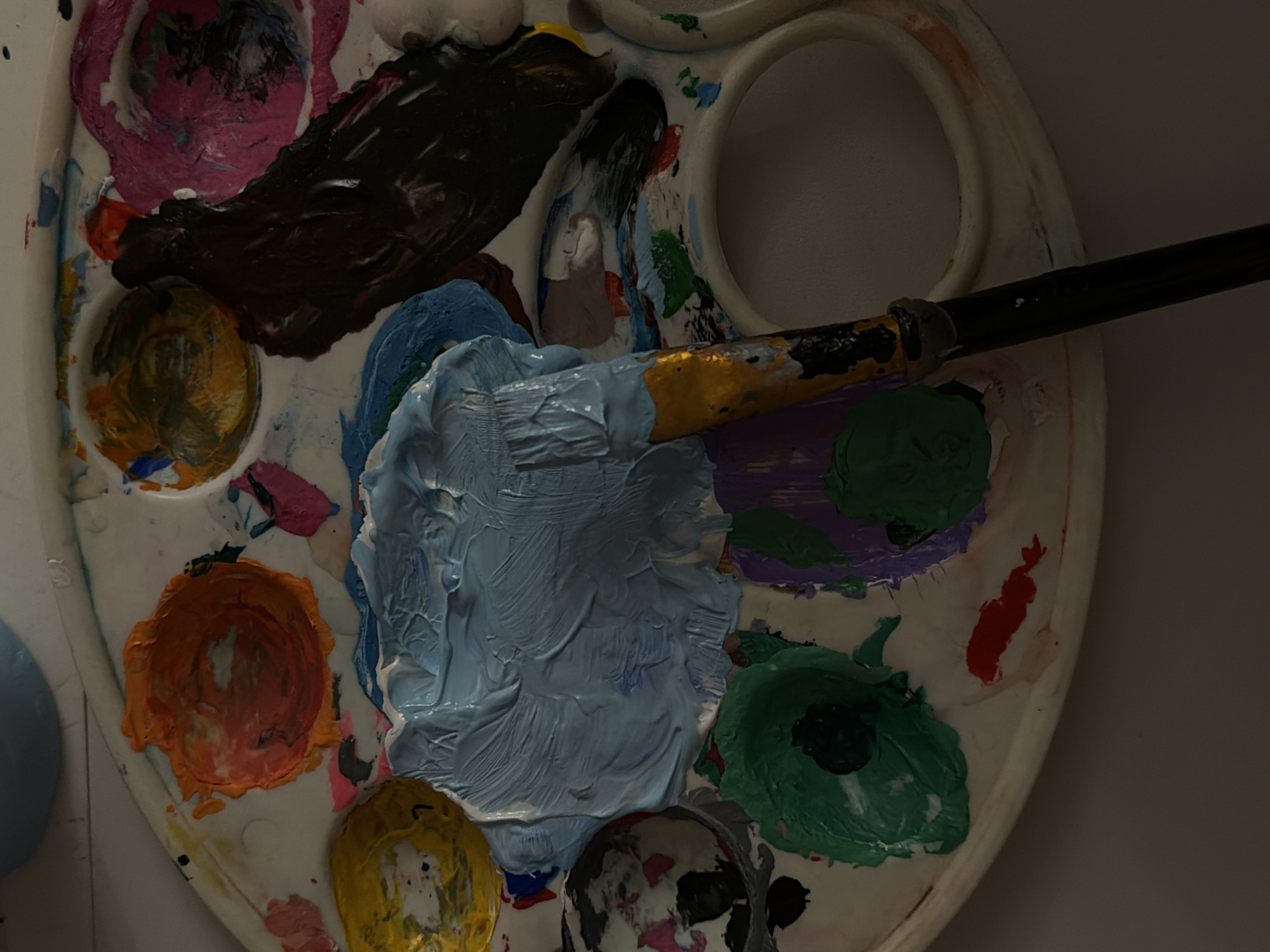 A top-down view shows a circular white plastic artist's palette with several wells containing different colored paints. There are paints of pink, brown, yellow, orange, blue, purple, green, red, black and white, all mixed together. A paintbrush with a gold ferrule and a black handle is resting across the palette, with the bristles coated in light blue paint. The palette is set against a white or light-colored surface. The city of Perm, Russia is not discernible from the image.