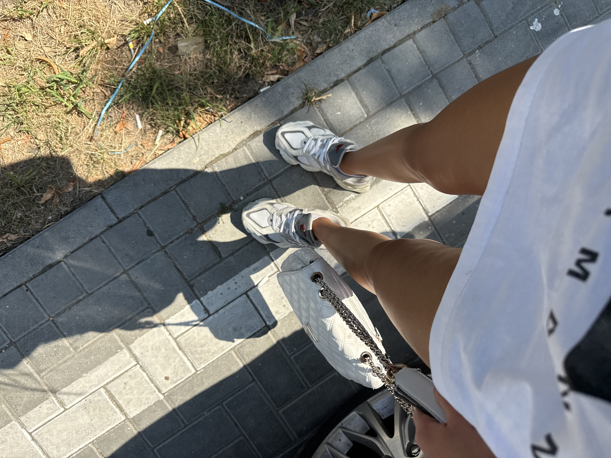 A first-person perspective shot captures a woman's legs and a white quilted handbag. She is standing on a sidewalk paved with rectangular gray stones, with dried grass and some litter visible to the right. Her legs are tanned and she is wearing white sneakers with grey accents and a white t-shirt. A shadow of the woman and her handbag is cast on the sidewalk. The image was taken outdoors on a sunny day, likely in Chișinău, Moldova.