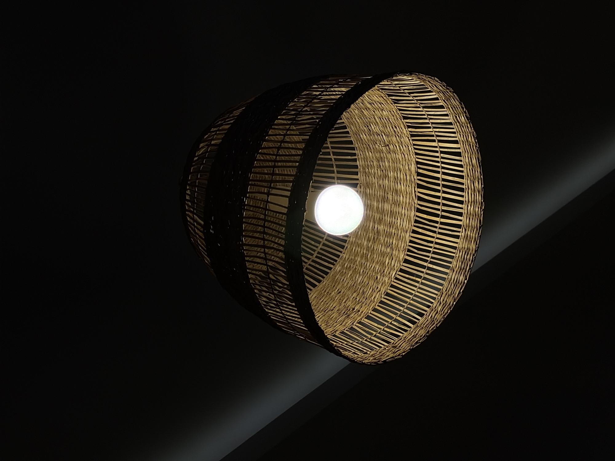A dimly lit, close-up shot features a cylindrical, woven lampshade with a lit bulb inside. The shade is constructed from a light-brown material, possibly rattan or wicker, arranged in horizontal bands with open spaces between. The bulb emits a bright, warm light. The background is mostly black, with a faint, angled light beam or shadow suggesting a ceiling or wall in the upper-right corner. The location is not evident from the provided image, and there is no information to suggest that it is in Budva, Montenegro.