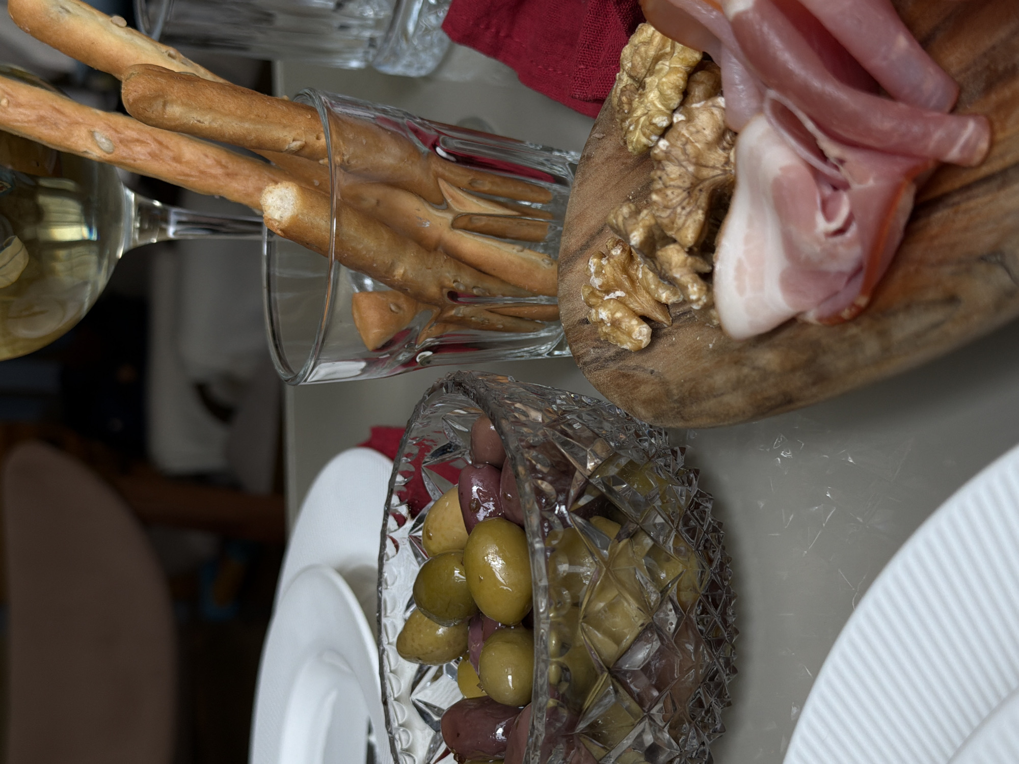 A top-down shot shows a table with various food items. A glass bowl filled with green and purple olives sits near two white plates. A wooden cutting board holds a pile of walnuts and slices of prosciutto. A glass filled with breadsticks rests near a wine glass. A red cloth is partially visible. The scene suggests a meal or snack setup on a table. There is no location information of Putilkovo, Russia available.