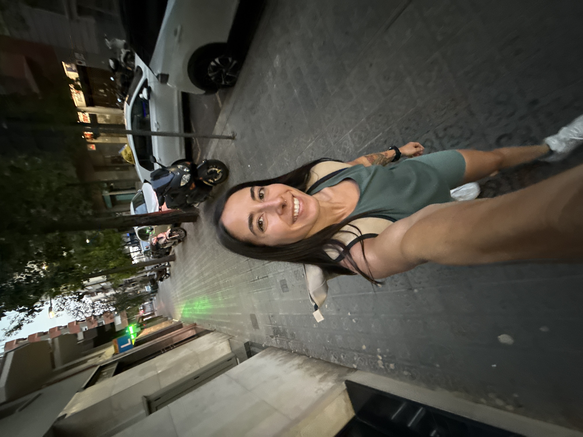 A woman with long dark hair and tattoos on her arm is taking a selfie on a city sidewalk in Barcelona, Spain. She is wearing a green tank top and shorts, and a backpack. The sidewalk is paved with grey stones, and there are trees and buildings on either side. Several motorcycles and cars are parked along the street. A green light is cast on the sidewalk from a sign on a building. The sky is dark, indicating it is either early morning or evening.