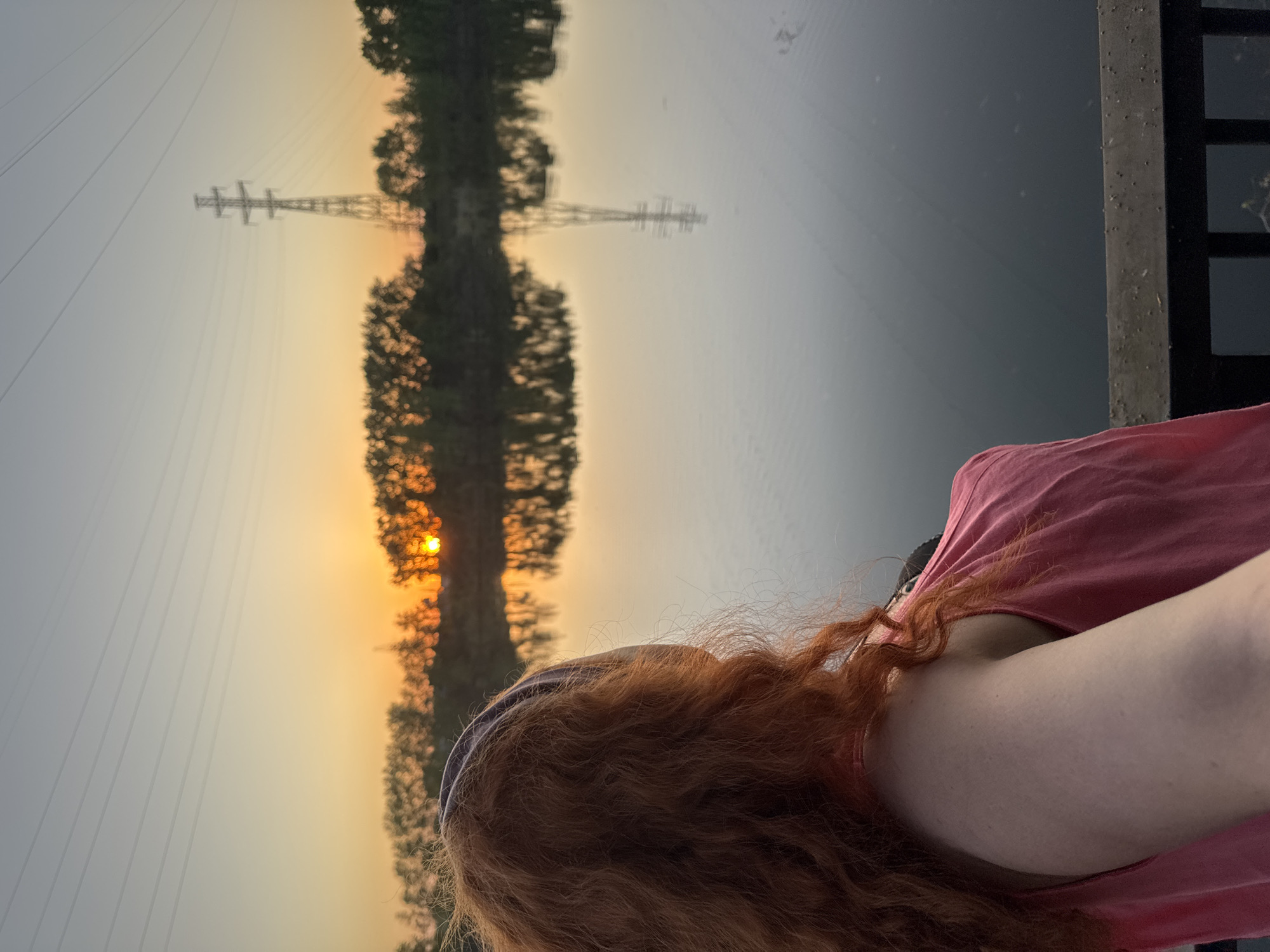The image shows a woman with long, wavy red hair standing near a body of water, likely a river or lake, in Chaikovsky, Russia. The photograph appears to be a selfie taken from a slightly low angle. The woman is wearing a pink t-shirt. In the background, across the water, is a reflection of a tree line and an electrical power transmission tower silhouetted against an orange-hued sunset sky. Power lines stretch from the tower. The water is a dark, grey color. A concrete or metal railing is visible on the right side of the frame, next to the woman.