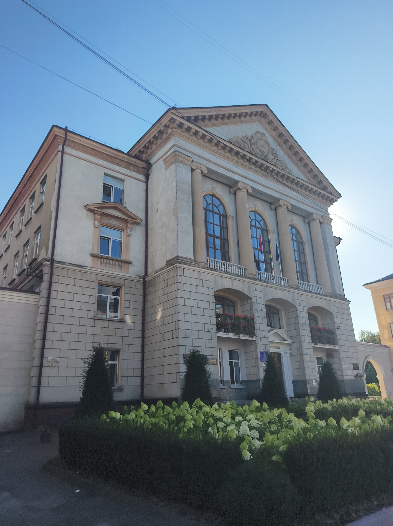 The image shows the Ternopil National Pedagogical University named after Volodymyr Hnatiuk in Ternopil, Ukraine. The university is a grand, multi-story building with classical architecture, featuring tall arched windows, columns, and decorative pediments. The facade is a light beige color with grey stone accents. In front of the building, there is a well-maintained garden with manicured hedges, conical evergreen trees, and abundant white and green flowers. The sky is a clear, bright blue. Power lines are visible in the upper part of the image. The lighting suggests it is daytime, with the sun casting a gentle glow on the building.