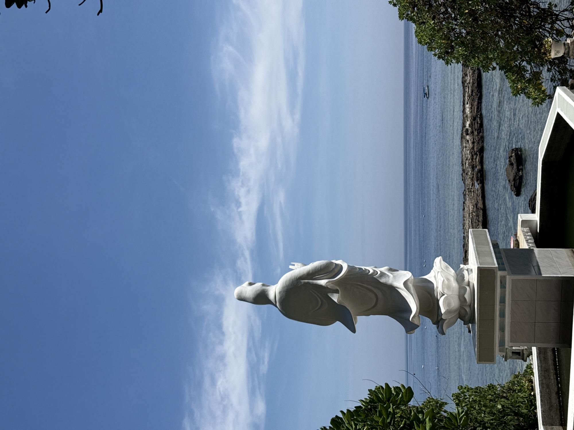 A full shot shows a white statue of a deity, likely Guan Yin, standing on a lotus pedestal against a bright blue sky with wispy white clouds. The statue is viewed from behind, with its hands held in a gesture of prayer or blessing. To the left and right of the statue, lush green foliage frames the scene. In the background, the calm, clear blue sea stretches to the horizon, dotted with a few small boats and rocky outcrops. The water is shallow enough in some areas to reveal the rocky seabed. The overall setting appears to be a coastal temple or spiritual site on Lý Sơn Island, a special administrative region in Vietnam, known for its volcanic landscapes and Buddhist temples. The sun is bright, casting soft shadows and highlighting the textures of the statue and the natural elements.