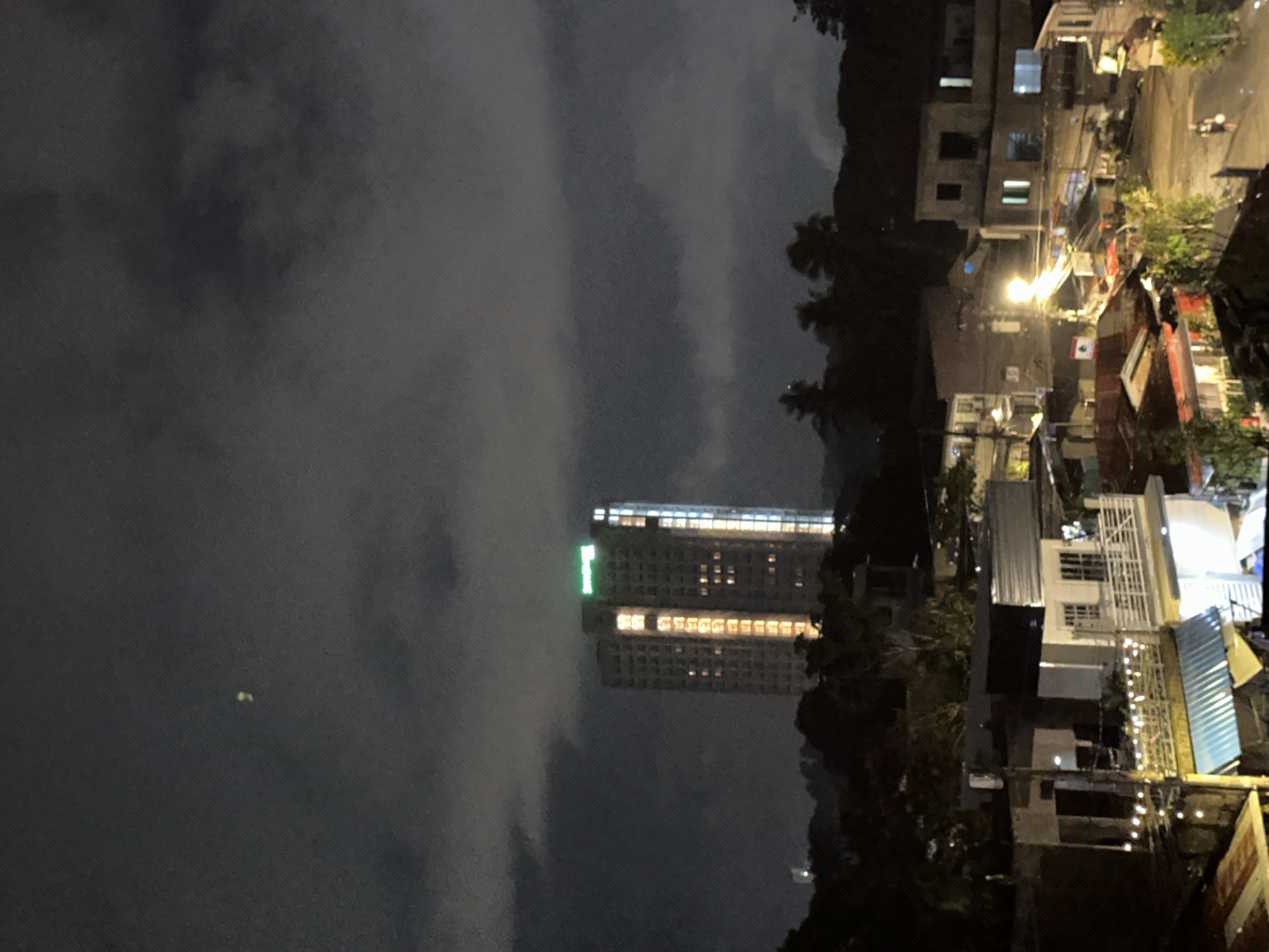 At night in Davao City, Philippines, a cityscape is observed. A tall building with illuminated windows and a green sign stands prominently in the middle ground, enveloped by fog and clouds. To the right, a neighborhood consists of various buildings, some lit with bright lights, surrounded by trees. A road winds through the neighborhood. The foreground contains a dark sky.