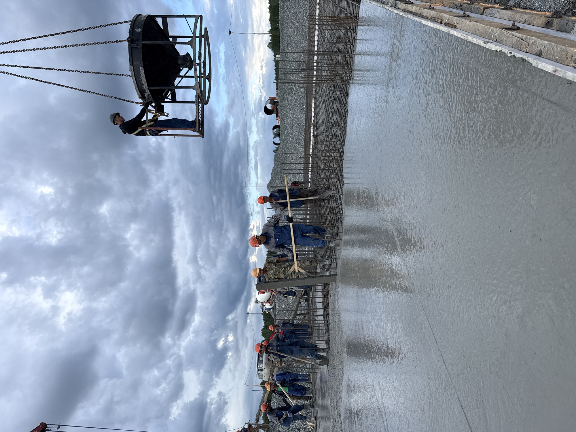 In Leninsk, Russia, construction workers are pouring concrete on a cloudy day. A group of men in hard hats and work clothes are spread across a rebar-reinforced surface, some using shovels and rakes to spread the wet concrete. A man in a harness stands on a platform attached to a large bucket suspended by chains, overseeing the concrete pour from above. In the background, there are piles of gravel, construction equipment, and distant trees under a heavily clouded sky. The overall scene depicts an active construction site in progress.