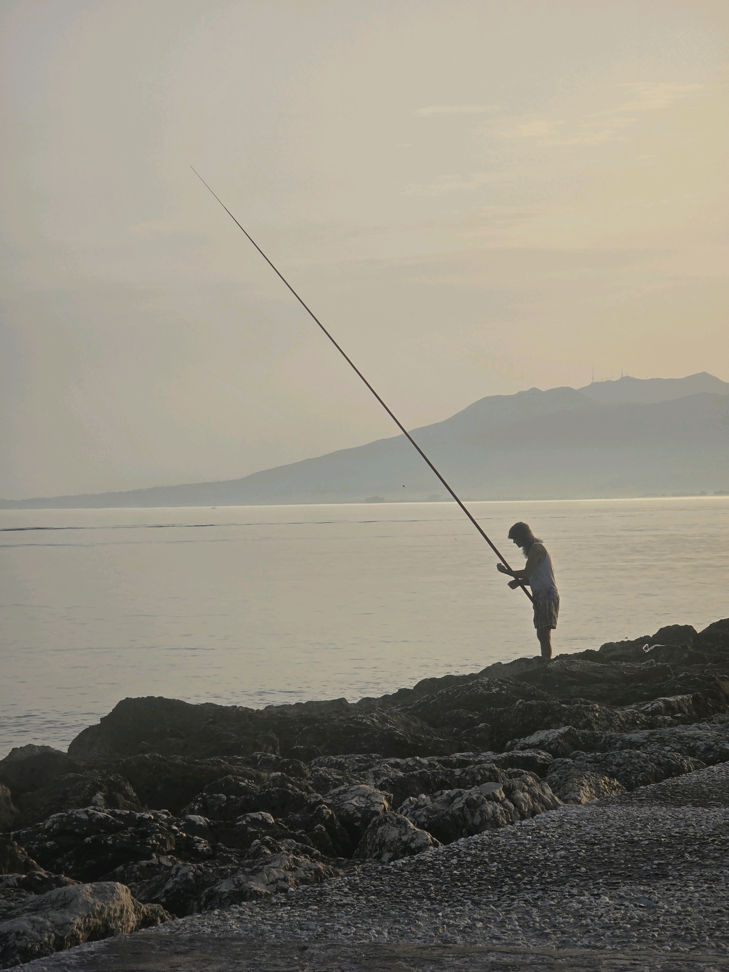 The image depicts a solitary individual fishing on a rocky shoreline, likely in Málaga, Spain, given the distinctive coastline and the presence of distant mountains, which could be the Montes de Málaga. The sky is overcast, creating a soft, diffused light that lends a serene atmosphere to the scene. The sea is calm, with gentle ripples reflecting the muted tones of the sky. The fisherman, silhouetted against the horizon, stands on a cluster of dark, rugged rocks, holding a long fishing rod that extends high into the sky. Their posture suggests focused concentration on the activity. In the background, a hazy mountain range stretches across the horizon, punctuated by what appear to be communication towers or structures. The shoreline is a mix of large, dark rocks and smaller pebbles, with a paved or gravel path leading towards the foreground. The overall impression is one of quiet solitude and communion with nature.