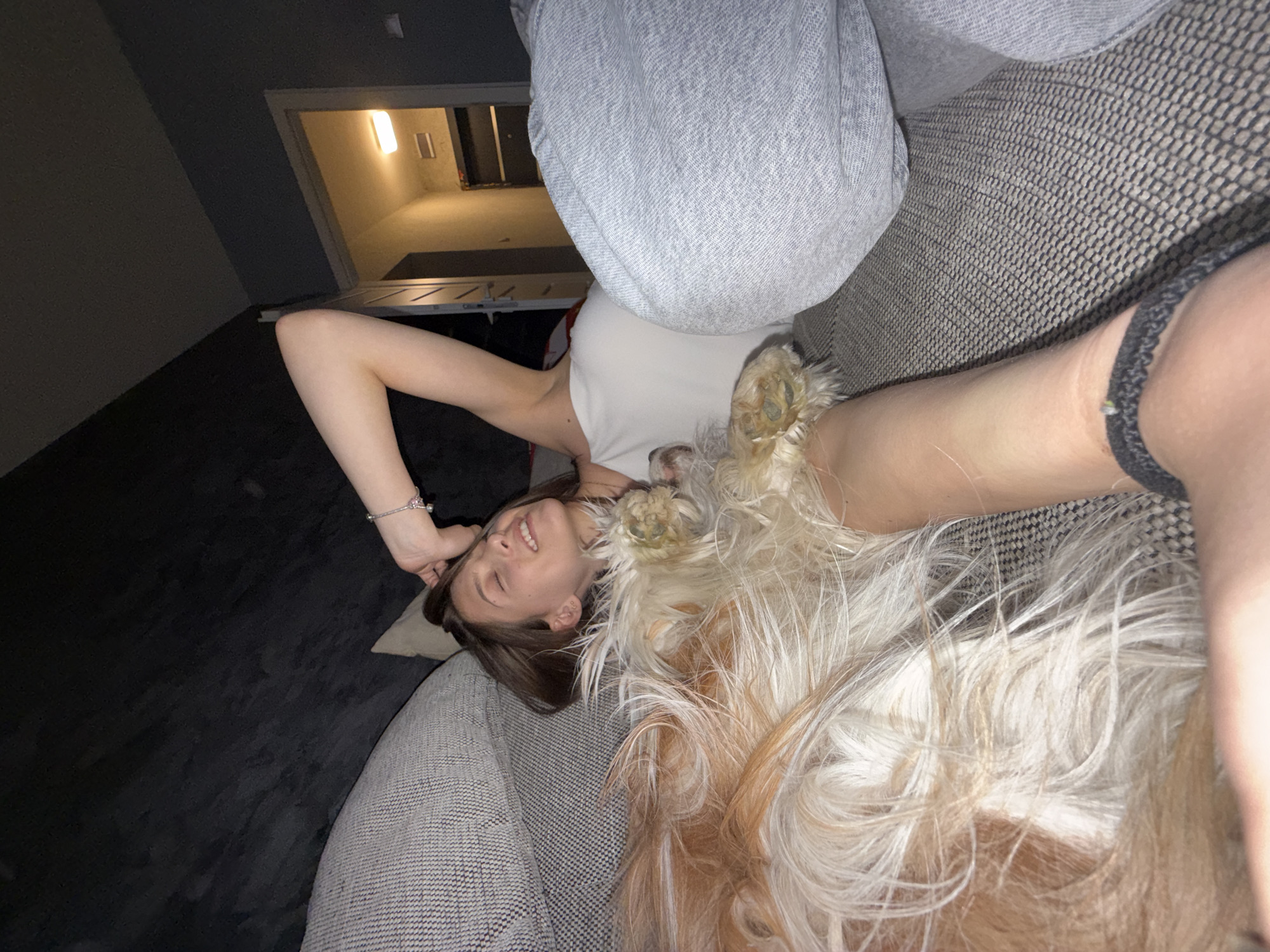 A young woman is lying on a gray sofa with a light-colored, long-haired dog lying on top of her. She is wearing a white tank top and gray sweatpants. The background includes a dimly lit room with a doorway leading to a brighter, possibly exterior, space. The location appears to be indoors, possibly in a residential building, although the city of Sofia, Bulgaria, cannot be confirmed from the image.
