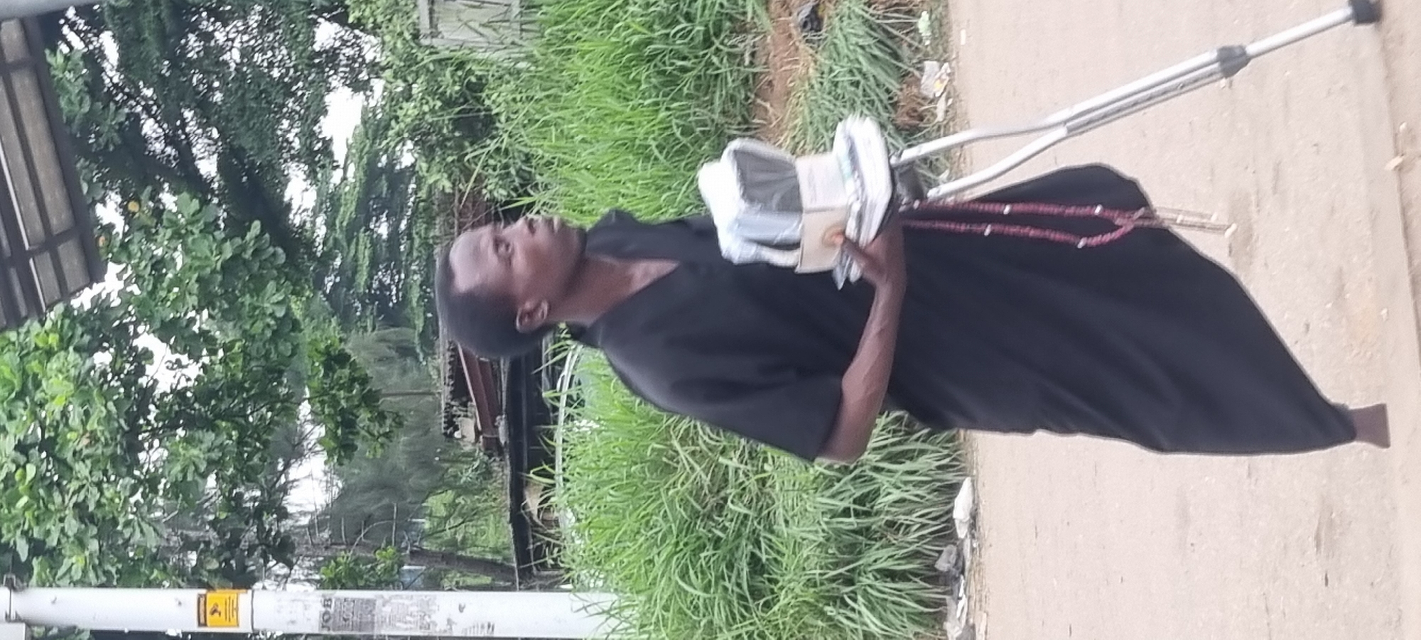 A medium shot captures a man in dark clothing standing with a crutch in Ikeja, Nigeria. The man, with dark skin and short hair, is looking to his right. He is holding a stack of items wrapped in plastic and paper, resting on a small box, in his left hand. A string of red beads with a metallic pendant hangs from his crutch. He is wearing a long, dark garment that covers his legs, and one bare foot is visible. The background consists of lush green grass, trees, and some distant structures that appear to be roofs or shelters, suggesting an outdoor, possibly urban or semi-urban, environment. The ground is a dusty, light-colored surface, likely a road or pathway. A utility pole with a yellow caution sign is visible on the left side of the frame.