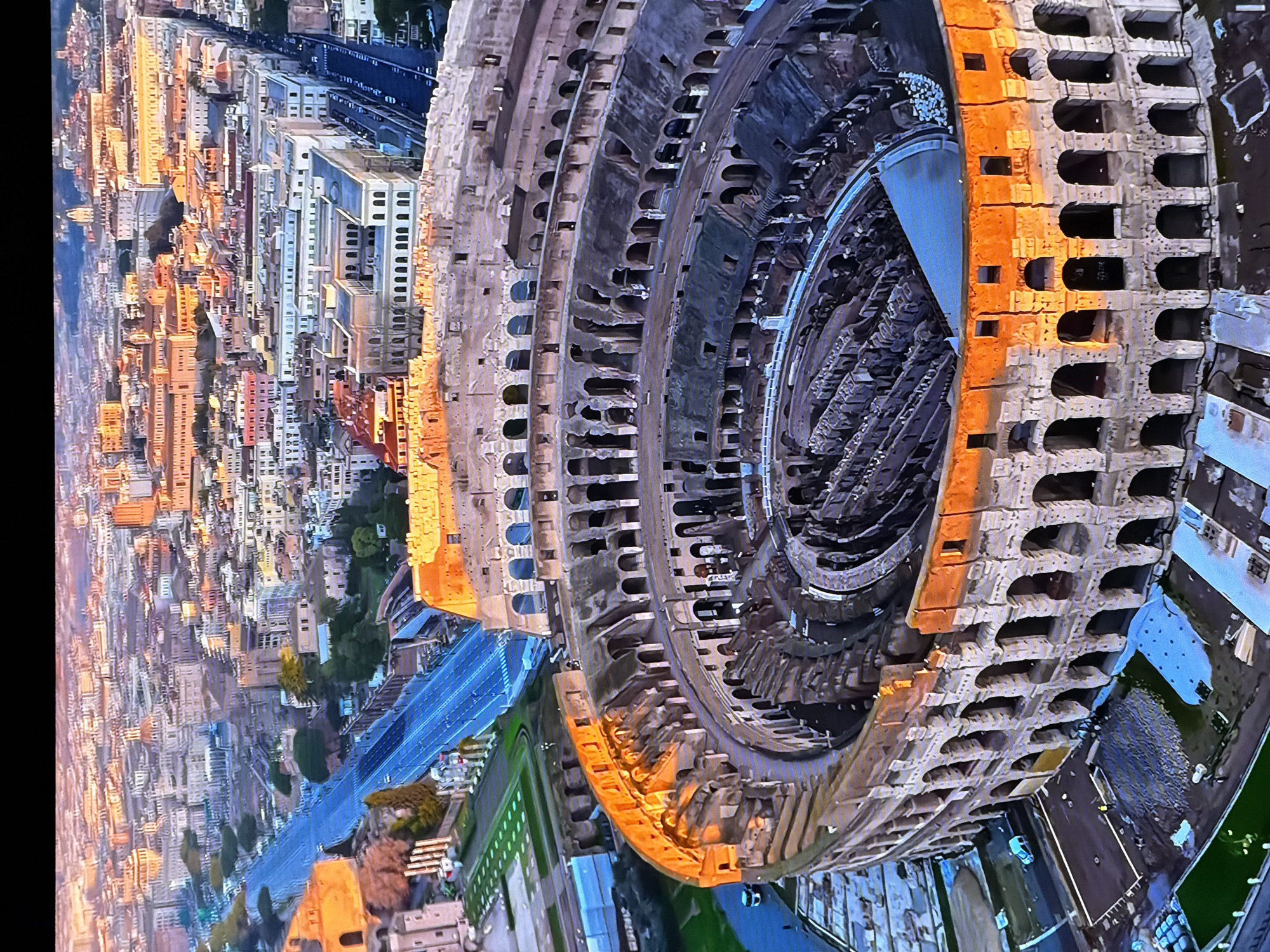This is an aerial view of the Colosseum in Rome, Italy. The ancient amphitheater is partially illuminated by the setting sun, casting a warm orange glow on its weathered stone. The vast structure is surrounded by the urban landscape of Rome, with buildings and streets stretching out into the distance. In the foreground, portions of the Colosseum's interior, including its tiered seating and central arena, are visible. The exterior walls, with their characteristic arches, stand testament to its historical significance. The image captures the grandeur of this iconic landmark against the backdrop of a vibrant city. The mention of Chirchik, Uzbekistan, is incorrect as the image clearly depicts the Colosseum in Rome.