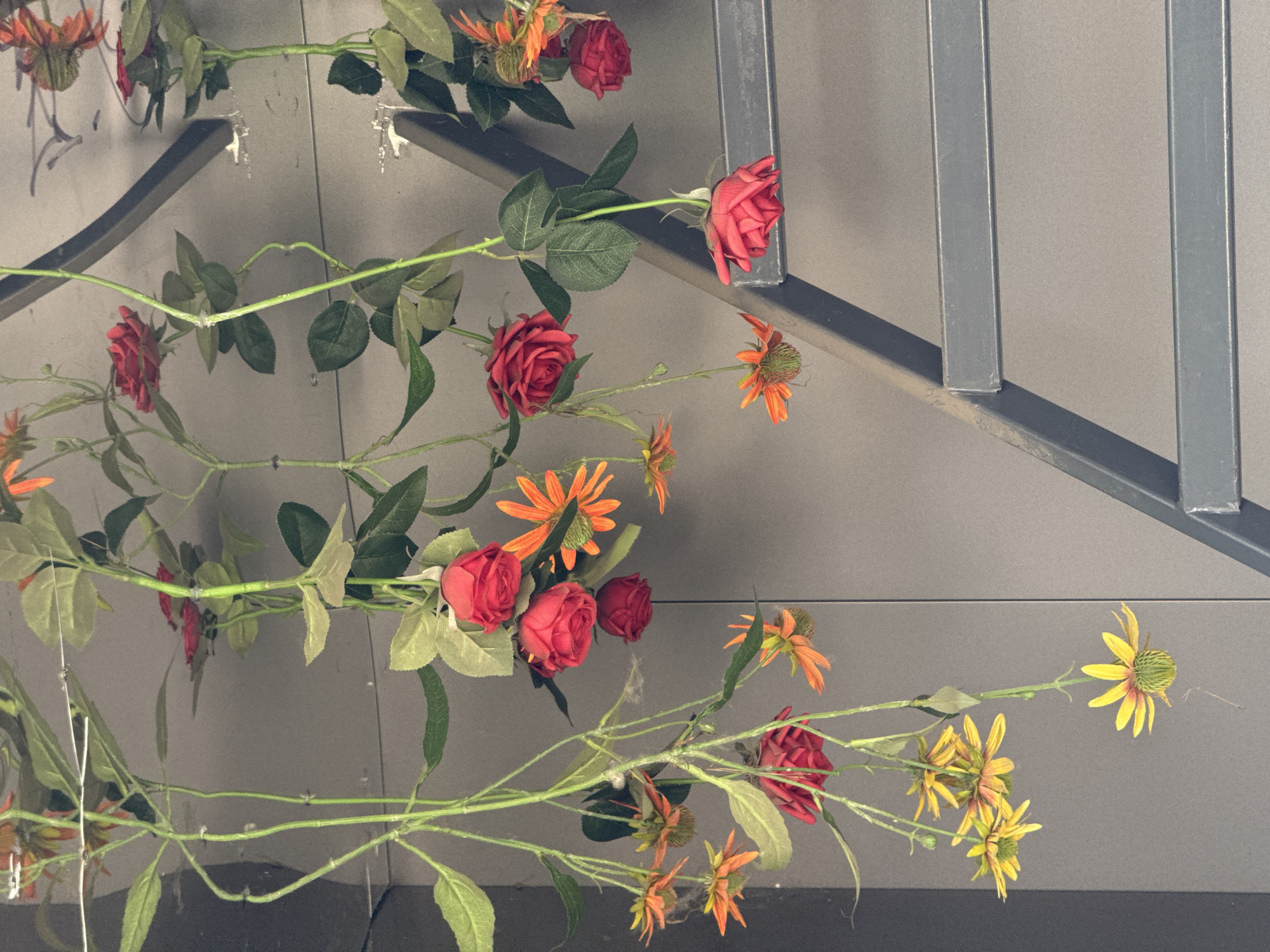 The image shows artificial flowers, predominantly red roses and yellow daisy-like flowers, hanging from an overhead support. The flowers are attached to green stems with artificial leaves. A grey, metal ladder structure is visible in the right half of the frame, leaning against a grey, textured wall. The lighting suggests an indoor environment, possibly with overhead lighting creating reflections. The image does not contain any people or visible signs indicating a specific location in Krasnoyarsk, Russia.