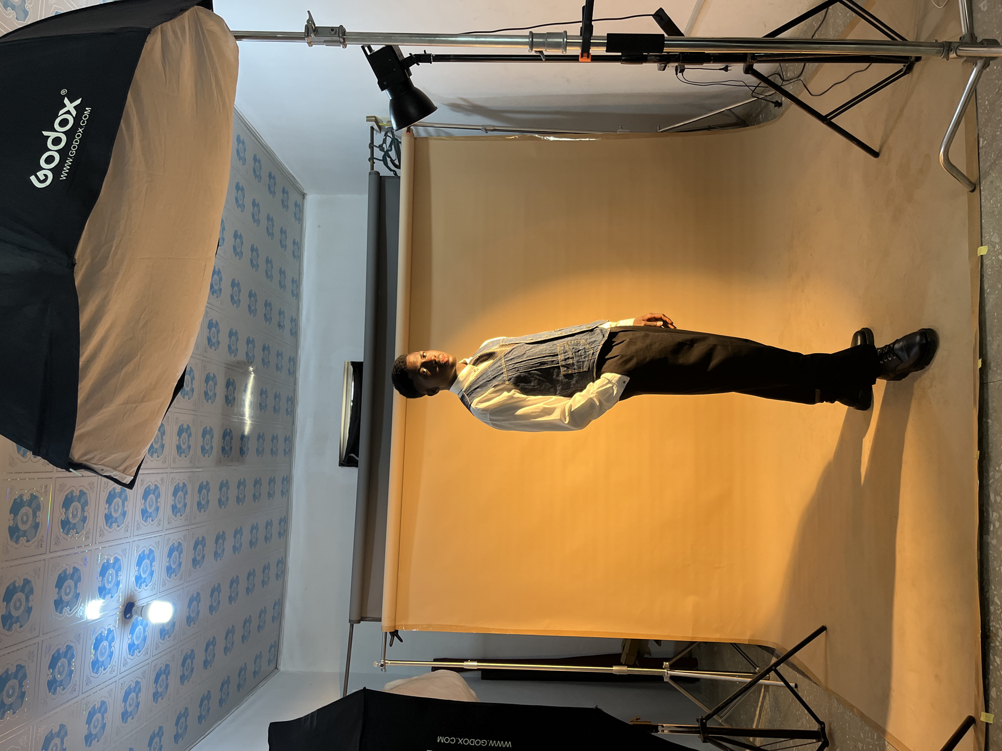 A man stands in front of a tan backdrop in a studio setting. He is wearing a white shirt, a denim vest, and black pants with black shoes. Overhead lighting equipment and two large Godox softboxes flank the scene. The wall to the left has a blue patterned tile. The backdrop appears to be supported by a stand, with lighting equipment visible at the top. The location is a studio, likely in Gura Top, Nigeria.