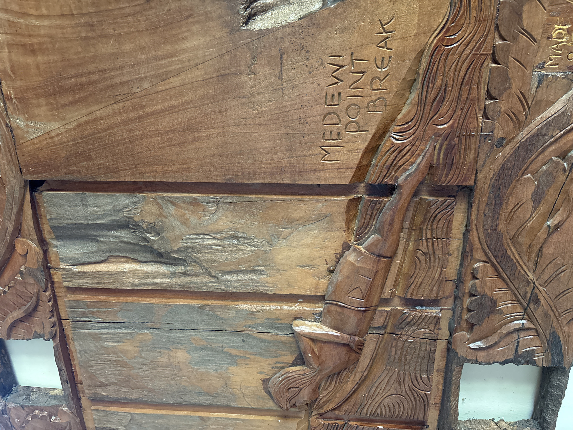 The image depicts a section of a carved wooden panel, possibly a sign or decorative element. It features the inscription "MEDEWI POINT BREAK," indicating the location is likely Medewi, a surfing spot near Pekutatan, Indonesia. The carving incorporates detailed imagery of a figure, possibly a surfer, with flowing lines suggesting water or waves. The wood has a warm, brown tone with visible grain and textures, some panels are aged or discolored. Some sections appear unfinished or broken. The overall impression is of a handmade, local artifact.