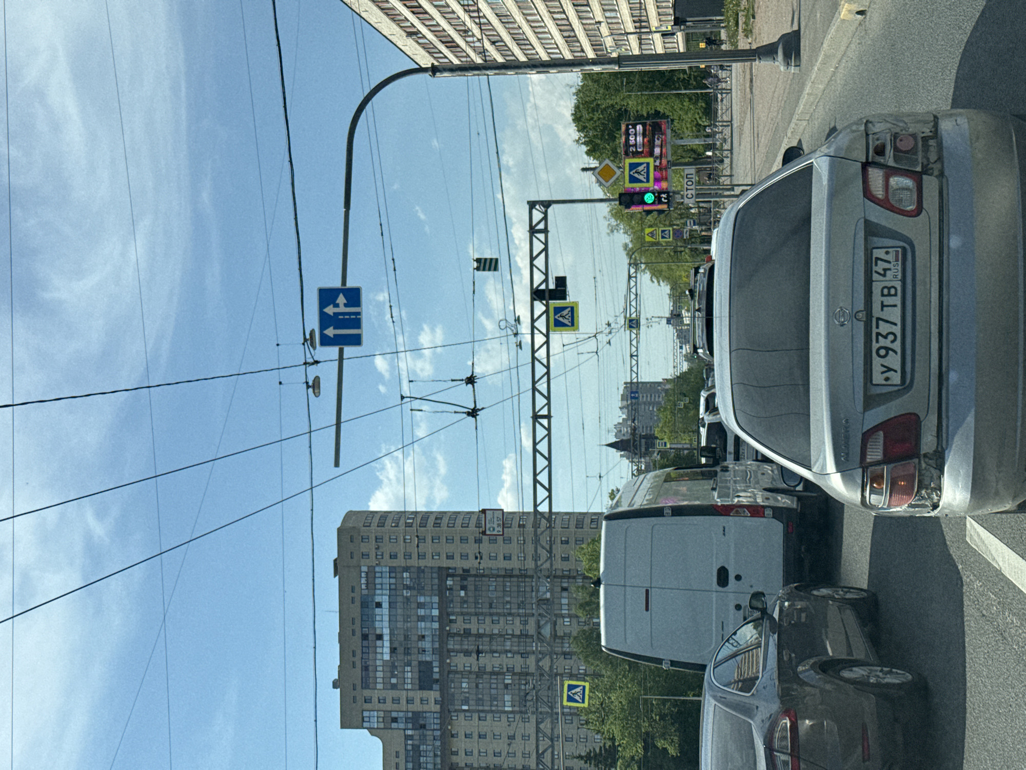 The image depicts a street scene in Saint Petersburg, Russia. The view is from inside a vehicle, likely a car, positioned in traffic. A silver car with a license plate reading "Y 937 TB 47" (registered in the Saint Petersburg area) is directly in front of the camera's POV. A white van is visible in the lane to the left. Several traffic lights are visible in the distance, with at least one showing a green light. Pedestrian crossing signs are present above the street. Overhead power lines stretch across the sky. High-rise buildings are on both sides of the road. The sky is partly cloudy. A blue road sign indicating a direction of travel is visible on the left.