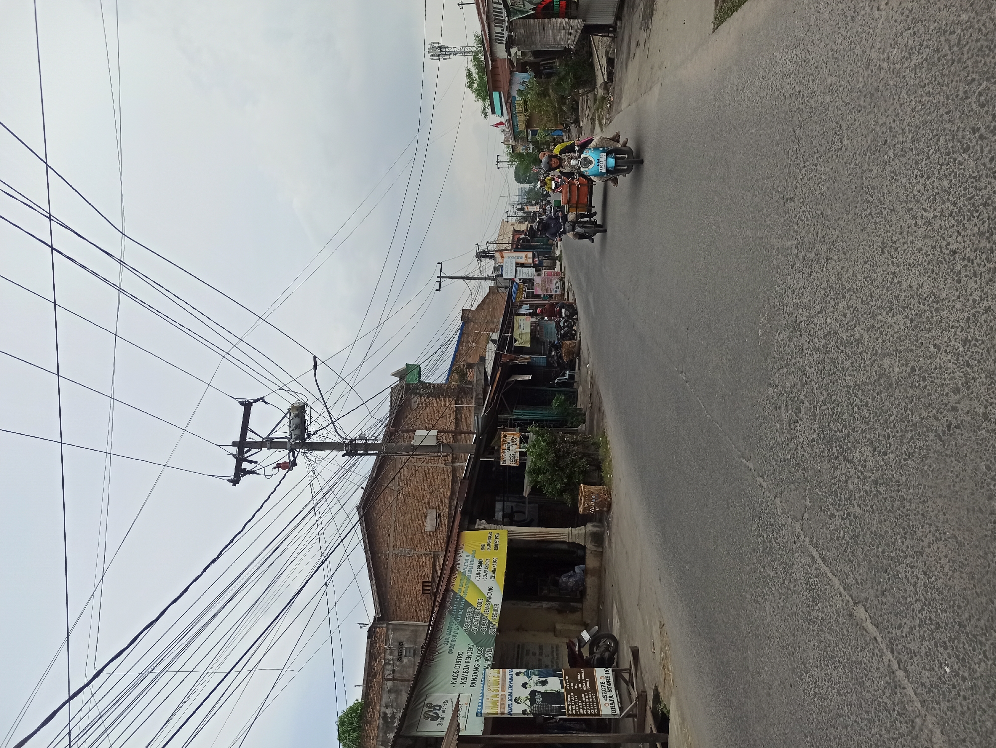 The image depicts a street scene in Medan, Indonesia, likely a commercial area judging by the storefronts lining the road. The sky is overcast, and numerous electrical wires crisscross overhead, attached to utility poles. On the left side of the street, there are low-rise buildings with awnings and signs, some of which appear to be shops selling clothing and other goods. A motorcycle is parked in front of one of the shops. The road is paved and appears to be in moderate condition with some cracks. Several people are visible riding motorcycles, with one motorcycle in the foreground carrying two individuals. In the distance, more buildings and utility poles are visible, suggesting a developed urban environment. The overall impression is of a typical day in a busy street in a Southeast Asian city.