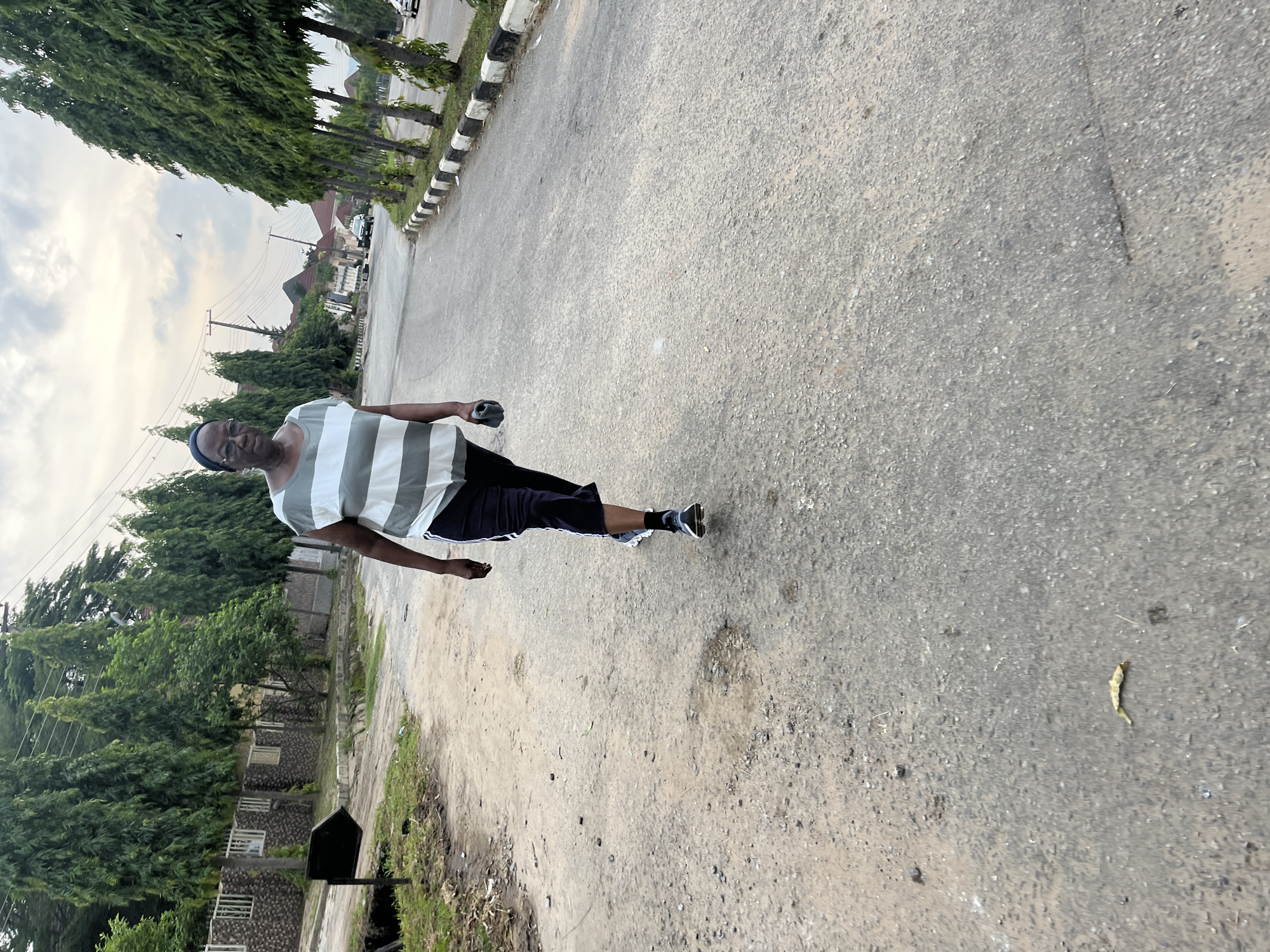 The image displays an outdoor scene featuring an individual walking on a paved road, captured in a portrait orientation that has been rotated sideways.
The main subject is an adult, positioned slightly right of the center, walking away from the camera but with their upper body turned towards it. They are wearing a light-colored t-shirt with horizontal grey and white stripes, dark shorts or capris, and dark sneakers with white socks. In their right hand, they hold a dark, possibly metallic or plastic, object. Their expression appears neutral.
The setting is a paved asphalt road, which occupies a significant portion of the foreground and midground. The road surface is grey and shows some signs of wear and tear, with small patches of dirt or debris. To the immediate right of the road, there is a black and white striped curb or barrier. Beyond this barrier, a dense line of tall, slender evergreen trees runs parallel to the road. On the left side of the road, there are unpaved areas with dirt, some sparse grass, and a few small structures or objects. More trees, including some with denser foliage, are visible on this side, partially obscuring residential-style buildings with stone facades in the background. Overhead power lines and utility poles are also present in the distance.
The sky is overcast with soft, diffused light, suggesting either an early morning or late afternoon on a cloudy day. There are no harsh shadows. The overall impression is a residential or suburban street scene, consistent with a location like Abuja, Nigeria. No visible text is present in the image. The person appears to be engaged in a casual walk or stroll.