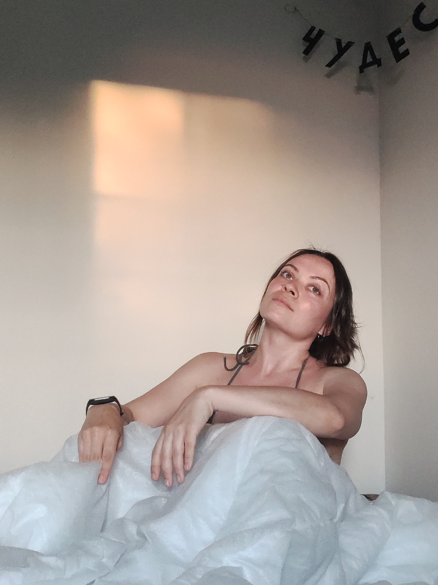 A woman with brown hair is sitting in bed, leaning on her hands. She is wearing a gray top and a black watch on her left wrist. The bed is covered with a white blanket. Behind her is a white wall with a rectangular patch of sunlight. At the top of the wall are black Cyrillic letters spelling out "ЧУДЕС". The location appears to be indoors, possibly a bedroom, in a city referred to as "Unknown, Unknown".