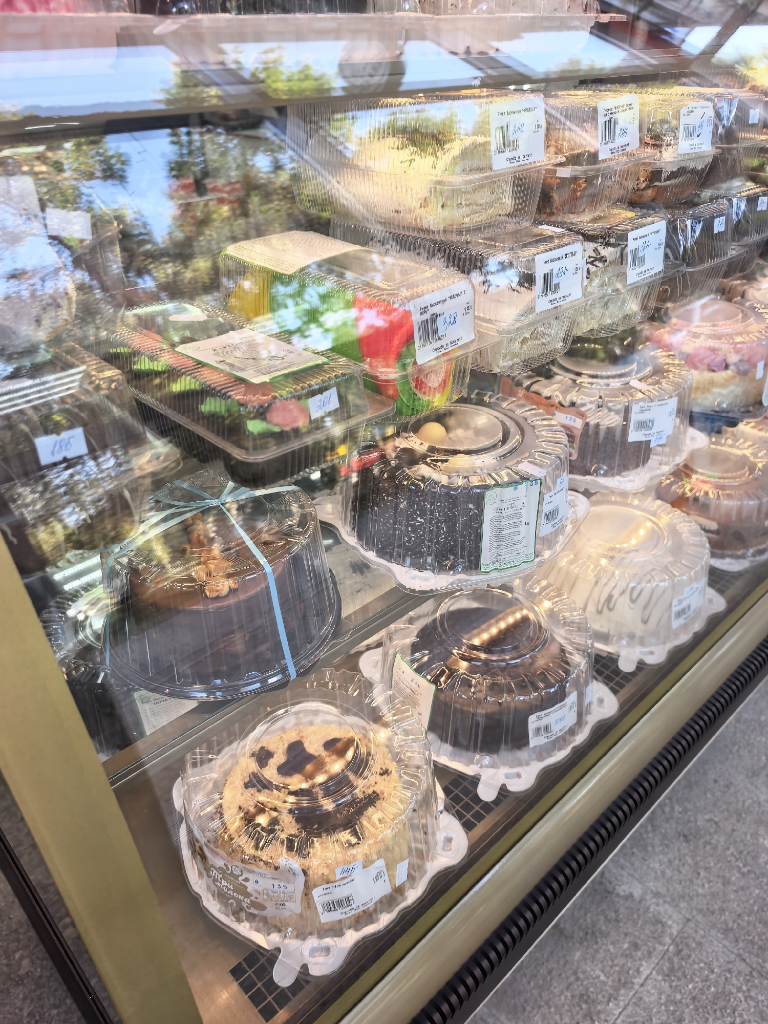 The image shows a display case filled with various cakes and pastries, likely in a bakery or grocery store. The cakes are in clear plastic containers, some with labels and prices visible. The display case appears to be made of glass and is well-lit, allowing for a clear view of the products. Reflections in the glass suggest an outdoor environment or a well-lit interior with windows. The overall impression is one of a retail setting where baked goods are being sold. The specific location in Sterlitamak, Russia, cannot be determined from the image alone.