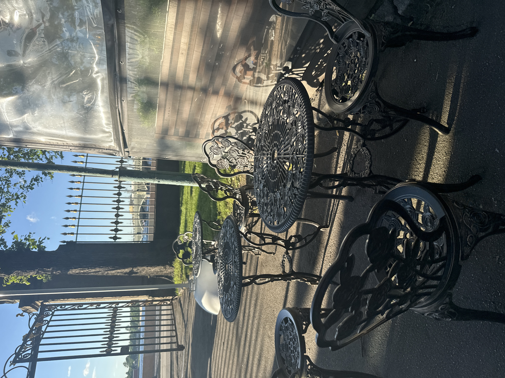 The image depicts an outdoor seating area with ornate, dark metal tables and chairs, possibly cast iron, arranged on an asphalt surface. The furniture features intricate floral designs. In the background, there is a tall tree with lush green leaves, a tall black metal fence with decorative gold finials, and a glimpse of a river or canal with a distant bridge. To the right, a transparent enclosure, likely for outdoor dining or a greenhouse, casts shadows and reflections. The scene is bathed in sunlight, creating strong contrasts and dappled shadows on the ground and furniture. The location appears to be a public space in Saint Petersburg, Russia, given the architectural style of the fence and the overall ambiance. There are no people visible in the image.