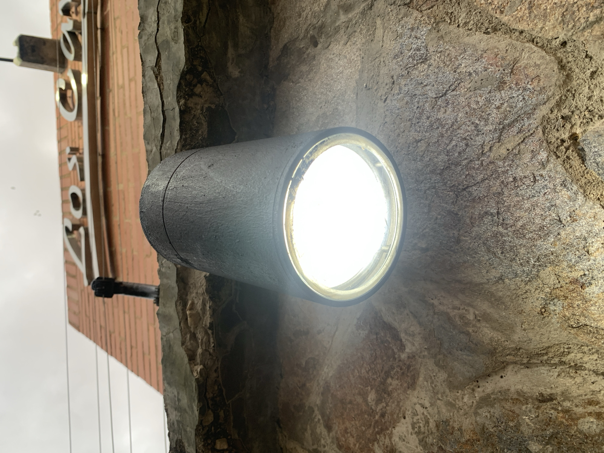 An eye-level, rotated close-up shot captures a bright, cylindrical black light fixture mounted on a rugged, natural stone wall. The powerful white light emitted from the fixture is intensely bright, illuminating the textured, variegated surface of the stone around it, which ranges in color from grey to earthy browns and reds.
To the left and above the light fixture, a brick building is partially visible, oriented ninety degrees, with neatly laid red bricks. A metallic, reflective sign with raised letters is affixed to this building, showing partial letters that appear to be "OS" or "SO", preceded by a curved element possibly from a "C" or "G". A black pipe or conduit extends from the brick wall. The transition between the brick building and the stone wall features cracked concrete or plaster.
The background reveals a bright, overcast sky, suggesting the photograph was taken during an overcast day, or potentially at dusk or dawn with the light fixture already active. The scene depicts an exterior architectural detail of a building in Pampatar, Venezuela, likely highlighting an entrance or pathway. There are no people present, and no specific activity is taking place.