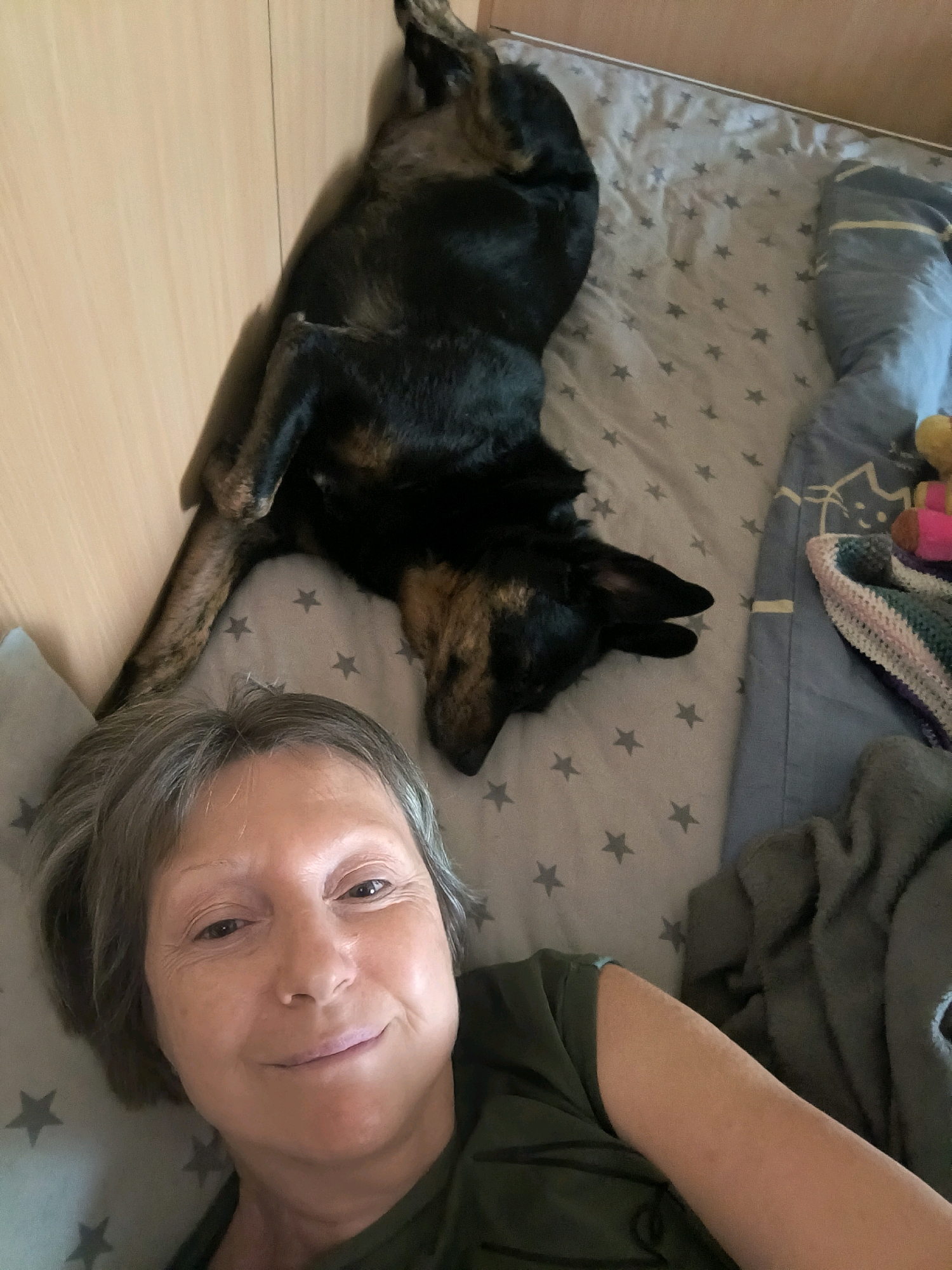 A person and a dog are lying on a bed. The person is in the foreground, looking at the camera and smiling. The dog is in the background, lying on its back with its legs in the air. The bed is covered with a grey sheet with star patterns. There are blankets and pillows on the bed. The environment appears to be indoors, likely a bedroom. There is no visible indication of the location being the city of Mykolaiv, Ukraine.