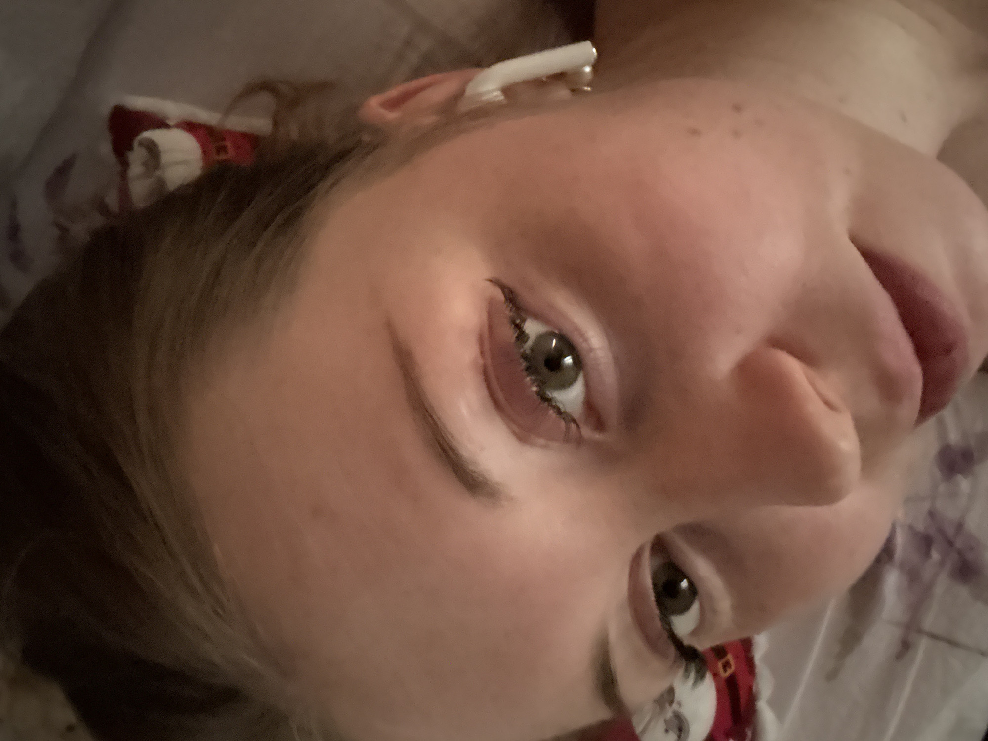The image depicts a close-up view of a person's face from a low angle. The person has fair skin, green eyes with black eyeliner, and brown hair. An earbud is visible in the person's left ear. A blurred red and white object is near the person's hair. The background includes a white and gray surface. The location is Unknown, Unknown.