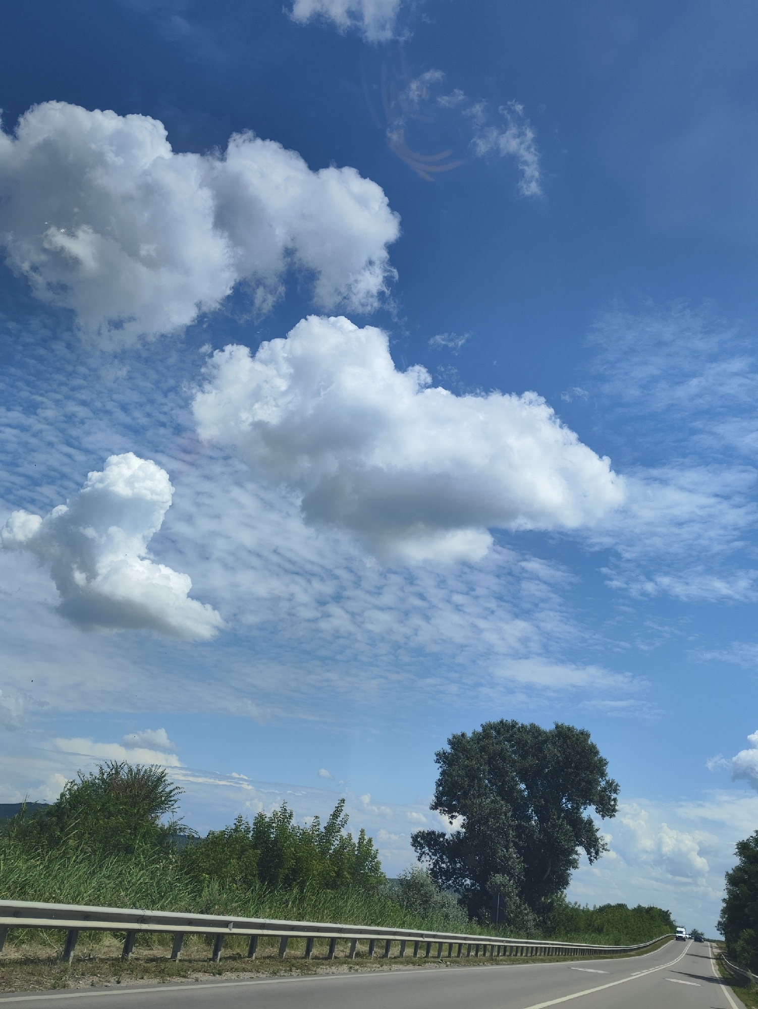 The image captures a scenic view of a road in Capriana, Moldova, on a sunny day. The sky is a vibrant blue, adorned with a mix of fluffy white cumulus clouds and wispy cirrus clouds. In the foreground, a paved road curves to the right, bordered by a metallic guardrail. Lush green vegetation, including tall grass, bushes, and a large, leafy tree, lines the side of the road. In the distance, a white vehicle is visible on the road. The overall impression is one of a peaceful drive through a natural landscape.