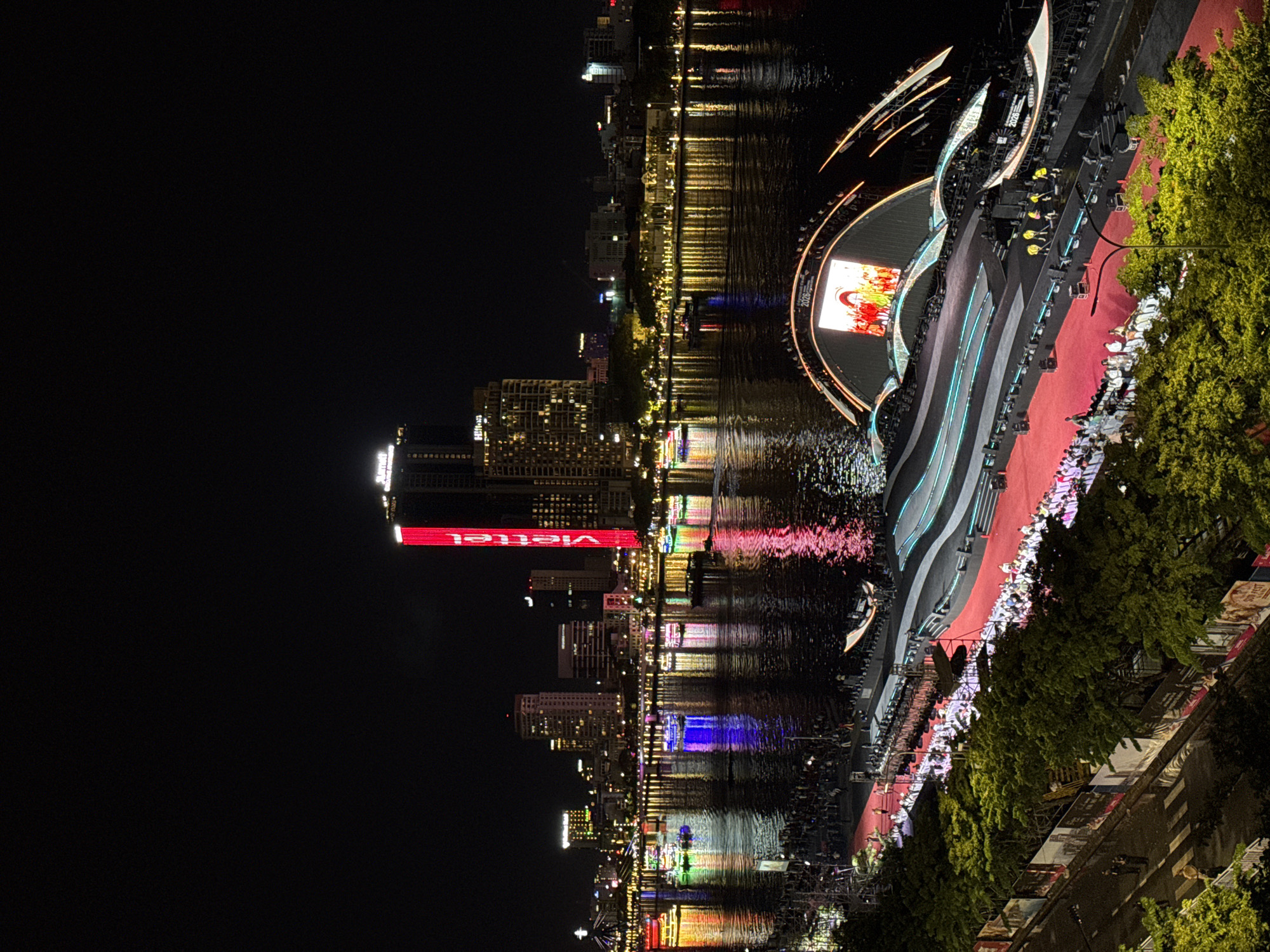 The image shows a nighttime aerial view of the Han River in Da Nang, Vietnam, during a festival. The river is filled with brightly lit boats, each adorned with colorful lights. On the riverbank, a large, elaborate stage is set up, featuring a massive arched screen displaying a lively performance. Red carpet leads up to the stage, and a crowd of people is visible in the foreground, seated or standing, observing the festivities. The city skyline, with its illuminated buildings and a prominent red "Viettel" sign, forms the backdrop. Trees and a street with a few people are also visible in the lower left portion of the frame.
