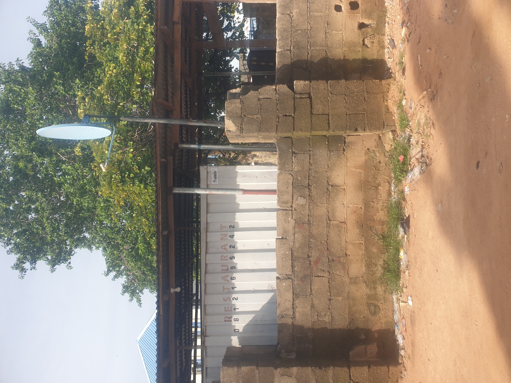 The image shows an outdoor scene in Jauro Yinu, Nigeria, featuring a brick wall in the foreground and a white container with "RESTAURANT" and a phone number spray-painted on it in the background. A satellite dish is mounted on a pole above the container, and a corrugated metal roof covers a structure behind the wall. Trees with green foliage are visible behind the structure, and the sky is overcast. The ground in front of the wall is sandy with some scattered debris.