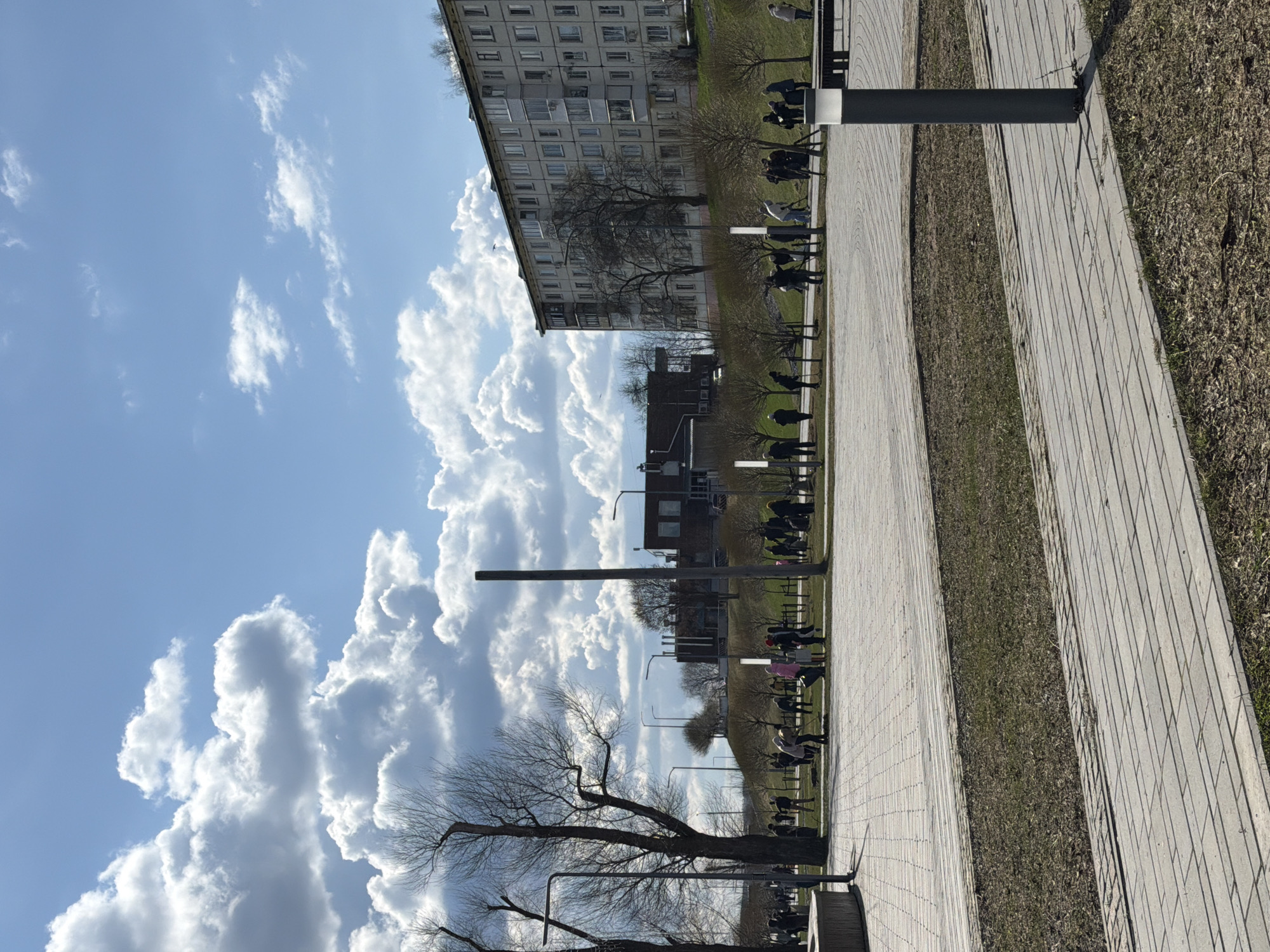 This image captures a bright, sunny outdoor scene, likely a public promenade or park in Votkinsk, Russia, during late winter or early spring. The photograph is taken from a slightly elevated perspective, looking across multiple parallel paved pathways separated by strips of dry, brownish grass.
In the background, several multi-story, light-colored apartment buildings with numerous windows, characteristic of post-Soviet architecture, define the urban setting. The sky is a vibrant blue, adorned with large, fluffy white cumulus clouds, indicating fair weather.
Numerous bare deciduous trees are scattered throughout the scene, their branches still devoid of leaves. Many people are visible in the middle distance and background, casually dressed and engaged in leisure activities. Some individuals are seated on benches along the paved paths, while others are standing or walking. Their forms are largely silhouetted against the brighter background. Several tall, dark poles, possibly streetlights or architectural features, are positioned along the walkways. The overall atmosphere appears calm and relaxed, with people enjoying the pleasant weather in a public recreational space. No visible text is present in the image.