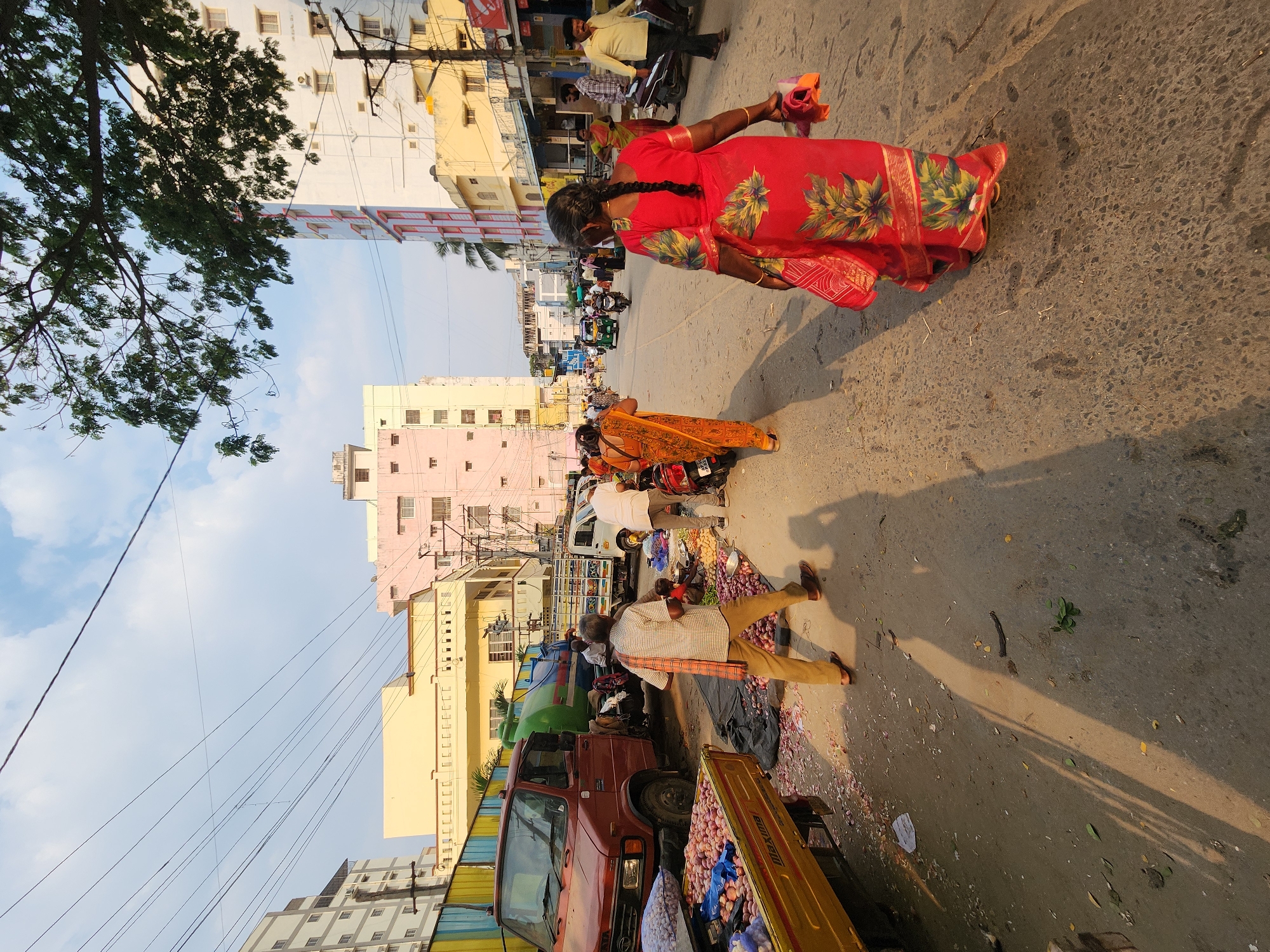 The image depicts a bustling street scene in Puttaparthi, India, likely a market area, under a clear sky with scattered clouds. The time of day is characterized by long shadows, indicating late afternoon or early morning.
In the immediate foreground, a person wearing a red sari with gold and green floral patterns walks away from the camera, carrying a small, wrapped item in their left hand. The paved road surface shows debris, including scattered onion skins and leaves. To the left, a portion of a yellow three-wheeled cargo vehicle, identified by "Maxima" branding, is visible, loaded with bags of red onions.
The midground features several individuals and vehicles. A man in a light-colored checkered shirt and light brown pants, with a striped cloth draped over his shoulder, walks towards the left foreground. To his right, another man in a white shirt and light pants stands near a ground display of vegetables, primarily red onions and potatoes, laid out on a dark tarp, suggesting a street vendor setup. Further left, a reddish-brown Tata truck with a blue and green tank in its bed is parked, partially obscured by a yellow construction barrier. A white pickup truck is visible behind the red truck. The street extends into the distance, populated by more pedestrians, parked motorcycles, and a green and yellow auto-rickshaw.
The background is dominated by multi-story urban architecture. Buildings of varying colors, including pale yellow, light pink, and white with red accents, line both sides of the street. Overhead power lines crisscross the upper portion of the frame against the sky. A large tree with green foliage is partially visible in the upper right, casting some shadow. Utility poles with numerous cables are also present.