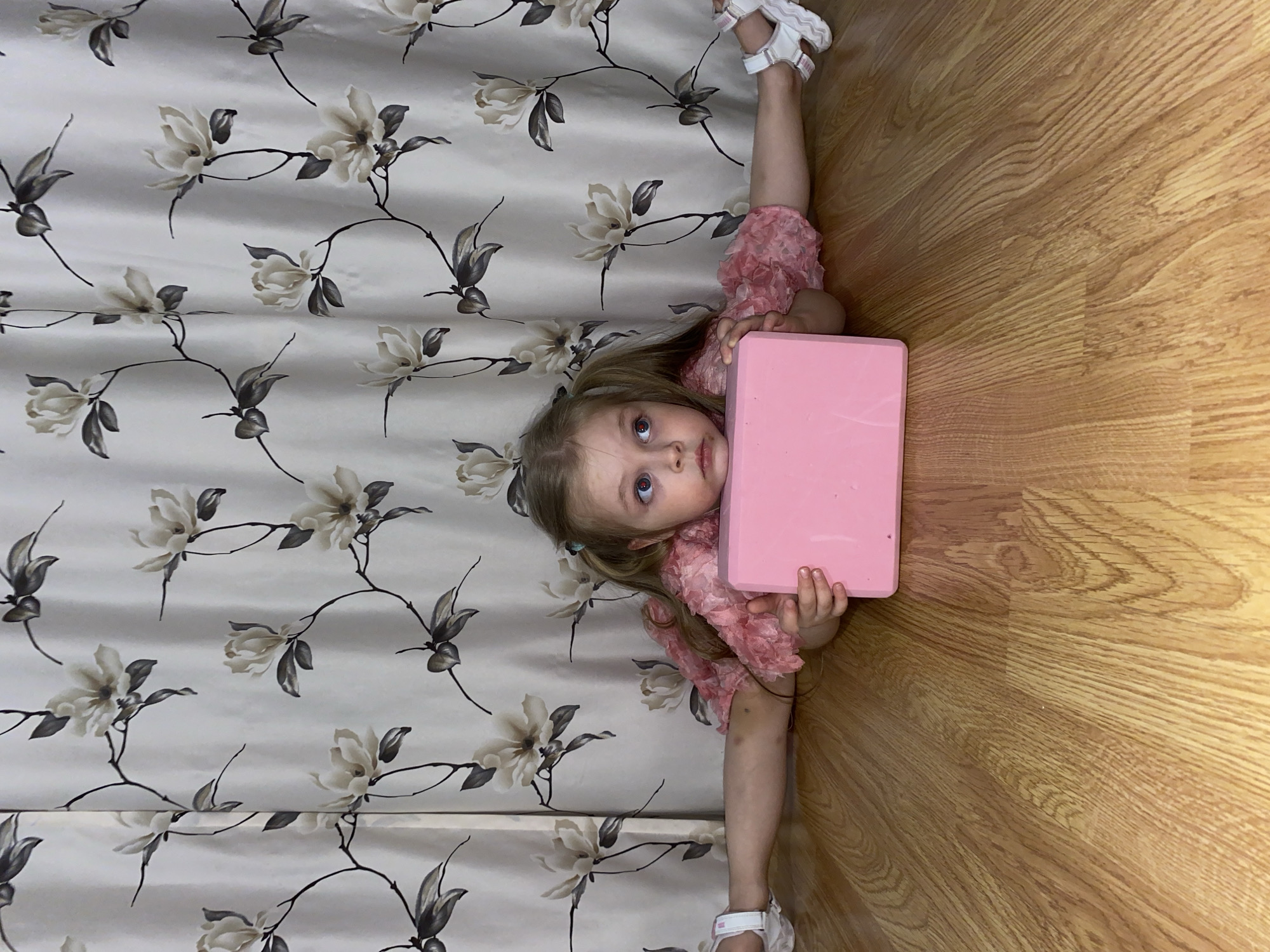 A small child, appearing to be a young girl with blonde hair, is positioned in the corner of a room in what appears to be a domestic setting. She is performing a split, with one leg extended towards a curtain or patterned wall, and the other leg extending towards a wooden floor. The child is holding a pink rectangular object, possibly a block, in front of her chest. She is wearing a pink, ruffled shirt and white sandals on her feet. The curtain or wall behind her features a floral pattern. The location is in Votkinsk, Russia.