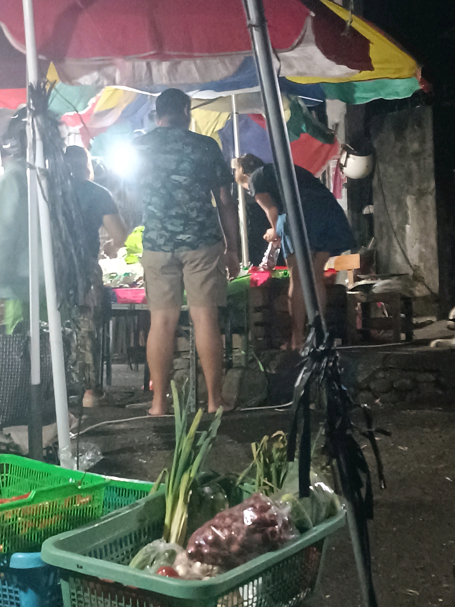 A dimly lit outdoor market scene is depicted, likely in Tabanan, Indonesia, under colorful awnings. Two individuals are visible. One, a person in a camouflage-patterned shirt and beige shorts, stands with their back to the camera, facing towards a table with various items. Another individual, wearing a dark blue dress, is also seen from behind, bending over and interacting with items on a table. In the foreground, two plastic baskets are filled with produce. The basket on the left is green and contains what appears to be vegetables and possibly red items. The basket on the right, also green, holds bundles of green onions, tomatoes, and items wrapped in plastic. A dark pole, possibly part of a market stall structure, runs vertically through the image, draped with black plastic strips. The ground appears to be dirt or pavement.
