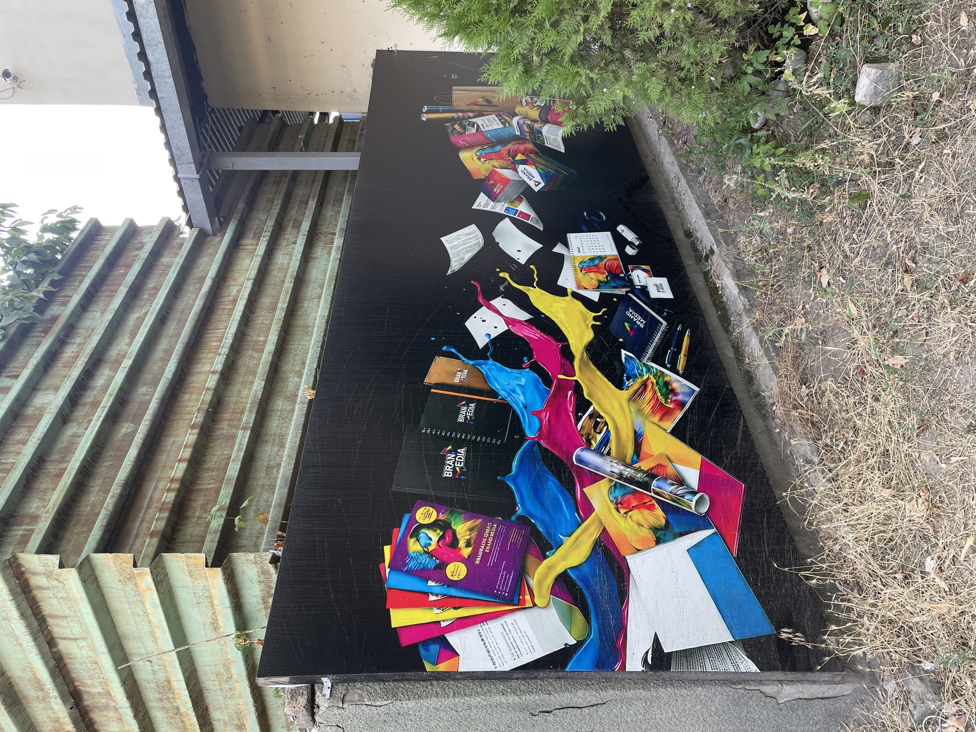 A mural depicting colorful splashes of ink and various print materials, including brochures, notebooks, and calendars, adorns a black billboard. The mural is located in Almaty, Kazakhstan, adjacent to a rusted corrugated metal fence and a light-colored building with a covered walkway. Dry grass and small plants surround the base of the billboard, indicating an outdoor urban environment. The scene is static and shows no human activity.