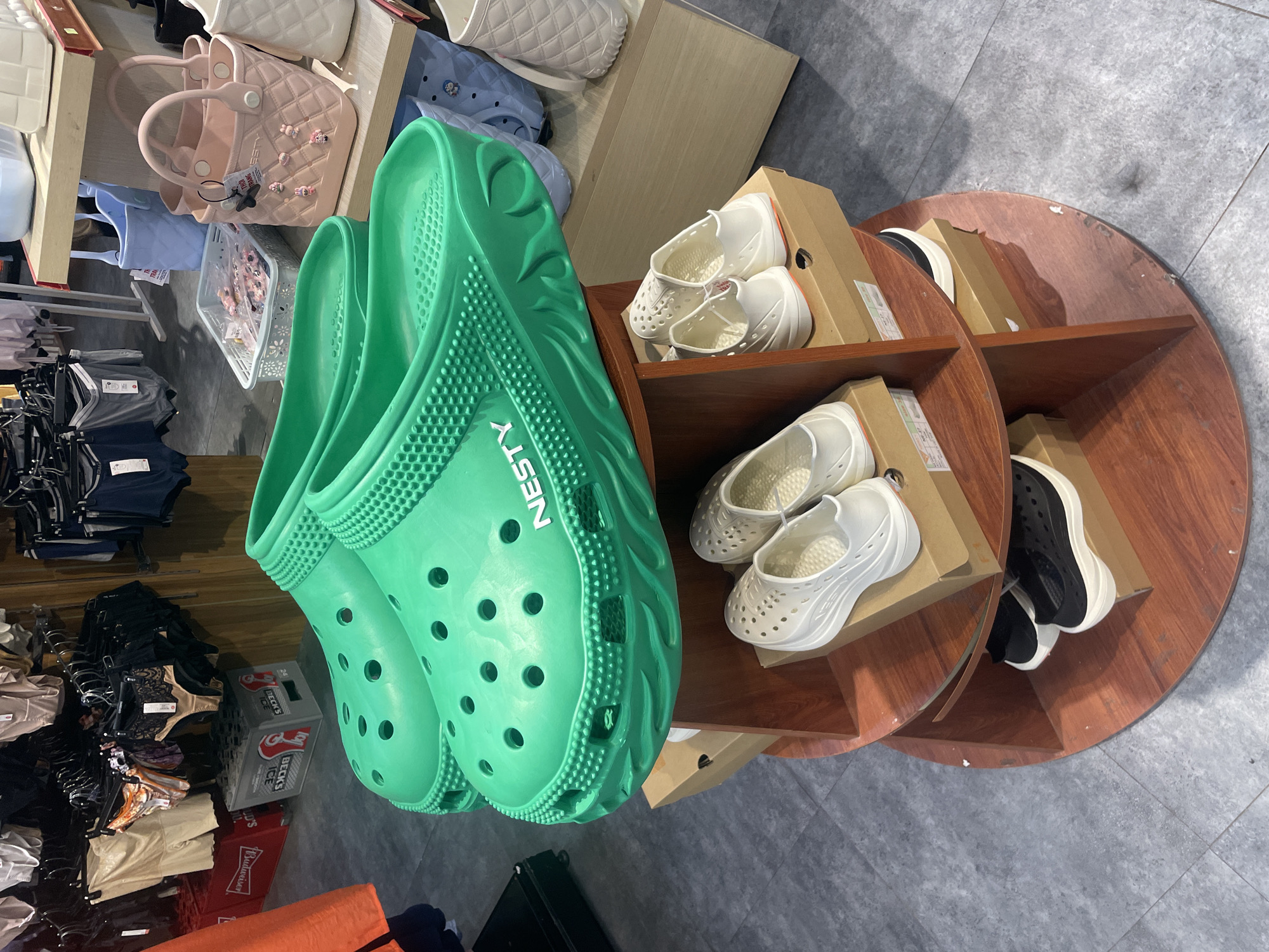 The image captures an overhead view of a retail display, likely within a store or market in Bac Nha Trang Ward, Vietnam. The main subject is a large pair of bright green, perforated clogs or slip-on shoes, positioned prominently in the foreground on the left. The word "NESTY" is visible in white letters on the side of these green shoes.
To the right, a round, multi-tiered wooden display stand holds several pairs of similar-style slip-on shoes in white and black, placed inside brown cardboard boxes. The white shoes feature perforated uppers and a slightly chunky sole.
In the background, shelves display various handbags, including a pink quilted handbag adorned with small charms and a light blue tote bag. Below these shelves, multiple clothing racks are filled with garments such as t-shirts, underwear, and other apparel. On the floor near the clothing racks, a grey crate with "BECKS ICE" written on it and a red crate with "Budweiser" are visible.
The floor is a grey tiled surface. The scene appears to be well-lit by artificial indoor lighting, suggesting daytime business hours. No people are visible, and the focus is on the displayed merchandise.