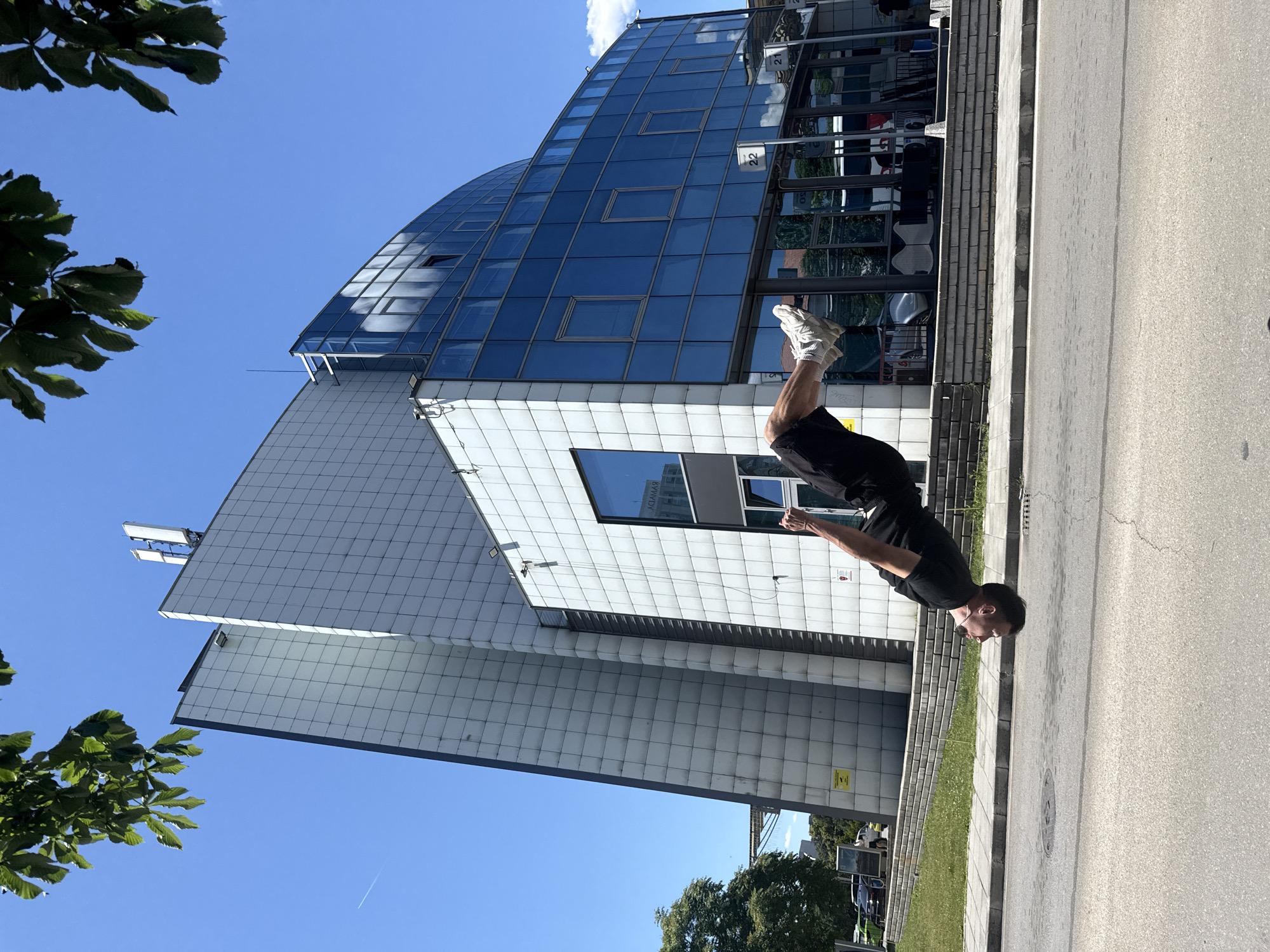 A man is performing a backflip in front of a modern, multi-story building with a glass facade in Sofia, Bulgaria. The building is angular and features white tiled sections and blue glass windows. The man is wearing a black t-shirt and black shorts, and he is captured in mid-air, upside down. The background shows a clear blue sky with some clouds, trees, and parked cars. The scene appears to be a public space, possibly a street or plaza, with a sidewalk and road in the foreground. The building's signage indicates it is located at numbers 21 and 22.