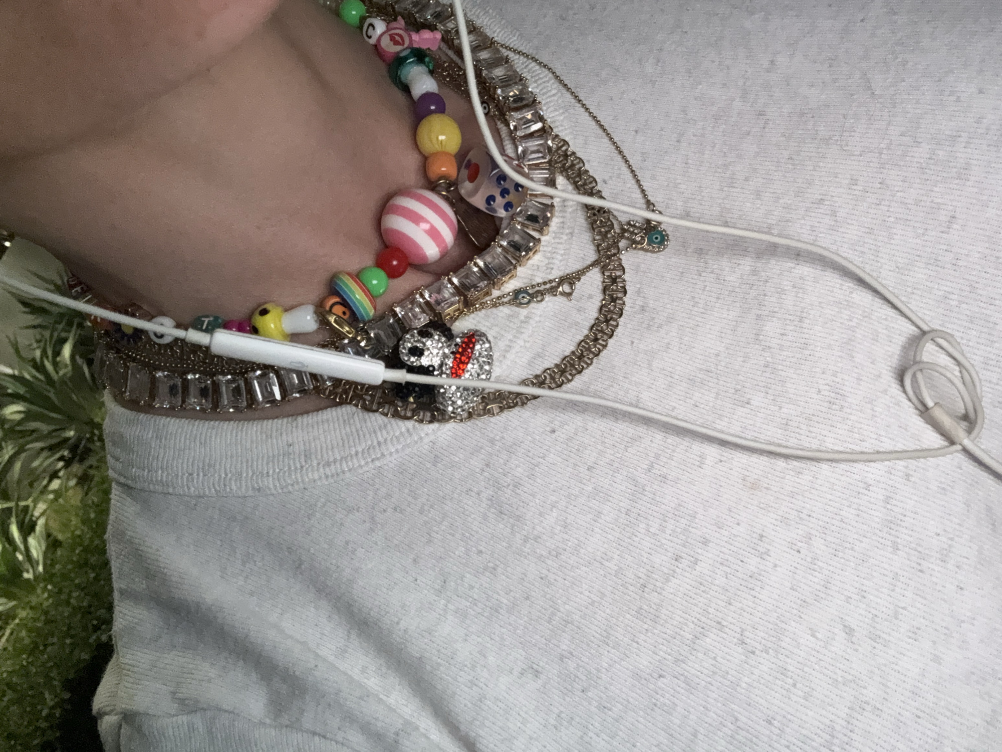 A close-up shot focuses on a person wearing multiple necklaces and a white t-shirt. The necklaces include a colorful beaded necklace, a chain with rhinestone-like settings, and other delicate chains. White earphones are draped over the necklaces. The background is blurred, showing some greenery.