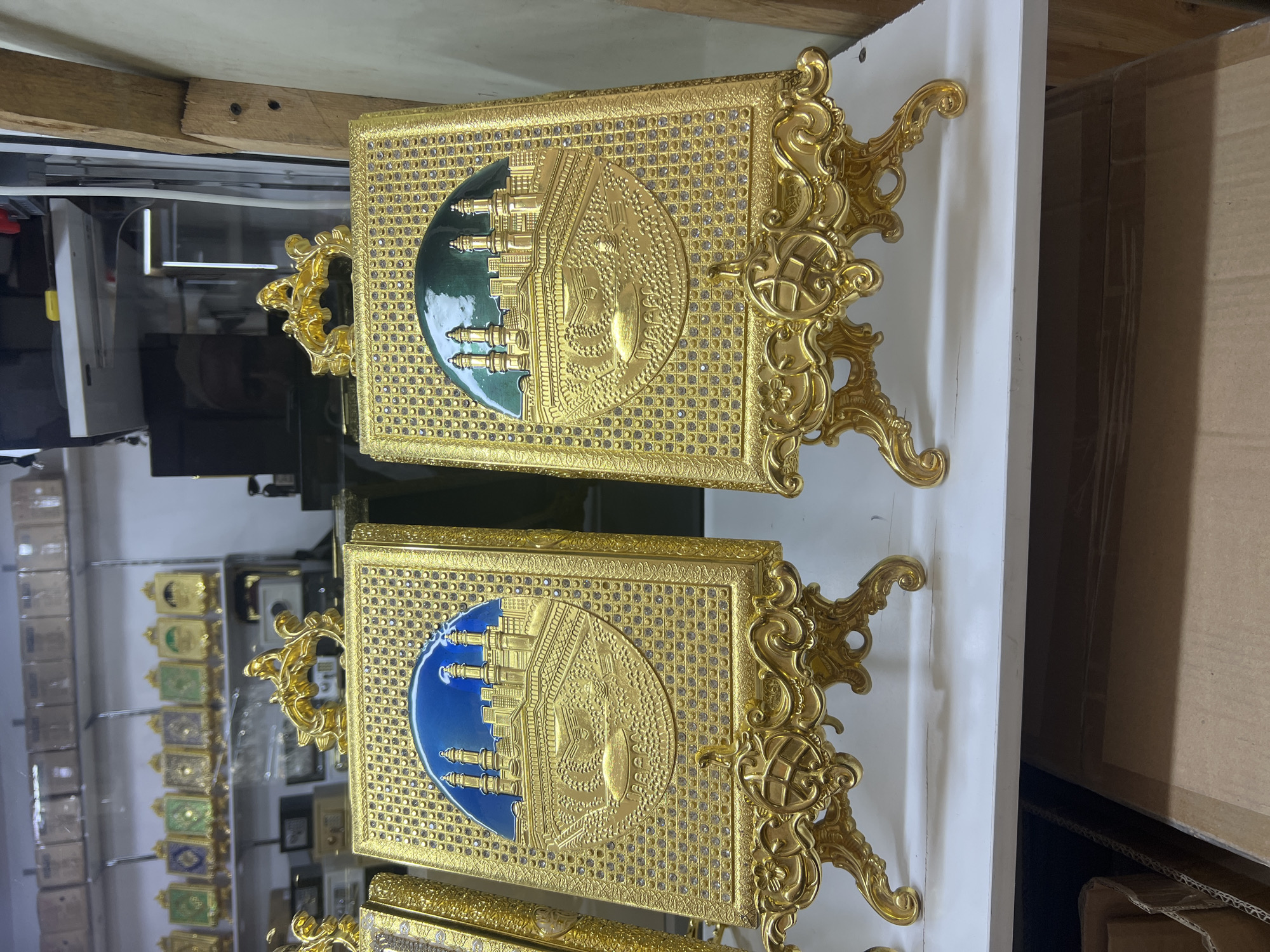 Two ornate, golden decorative plaques with Islamic motifs are displayed on golden stands on a white shelf. The plaque on the left features a raised, embossed image of a city skyline with minarets and a dome, set against a blue background within a circular frame. The plaque on the right has a similar raised, embossed image, but its circular frame displays a green background. Both plaques are decorated with numerous small, sparkling stones. Behind the plaques, on higher shelves, are rows of smaller decorative items. The shelves are part of a retail display, likely in a shop. The bottom of the image shows cardboard boxes stacked beneath the shelf. The context provided about Eshanguzar, Uzbekistan, is not directly observable from the image itself.
