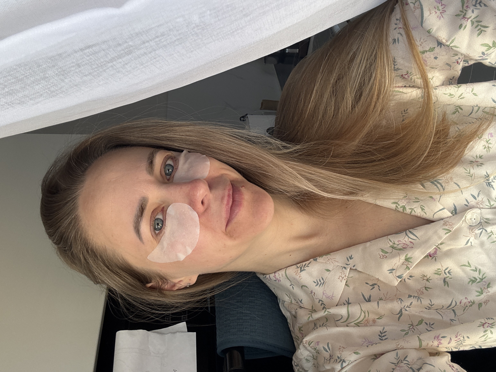 A woman is lying down with under-eye patches. She has light-colored hair and is wearing a floral pajama top. The background includes a white curtain and a dark object, possibly furniture. There is no evidence or visual clues to indicate this image was taken in Chaikovsky, Russia.