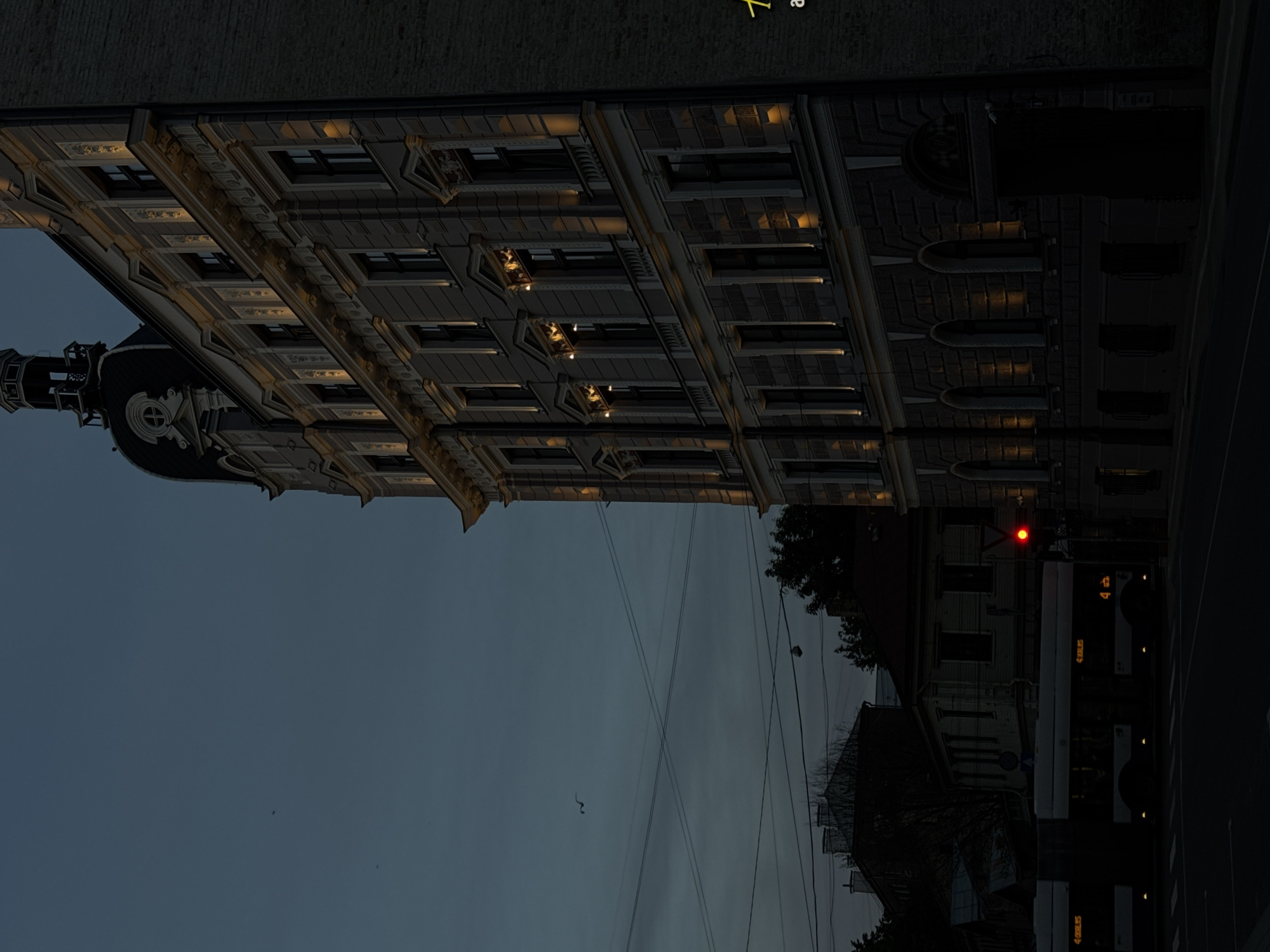 The image depicts a street scene in Riga, Latvia at dusk. An ornate, multi-story building with arched windows and decorative trim is illuminated by soft, yellow exterior lights. The building features a domed tower with a dark roof. In the foreground, a trolleybus, identified by the markings "4 KRLIS" on its side, is stopped at a traffic light that is red. Across the street, a row of darker, older buildings is visible. The sky is a deep, dusky blue with some cloud cover and power lines crisscrossing the view. A single bird is in flight in the upper left portion of the frame. The overall atmosphere is quiet and urban, with the lighting suggesting either early morning or late evening.
