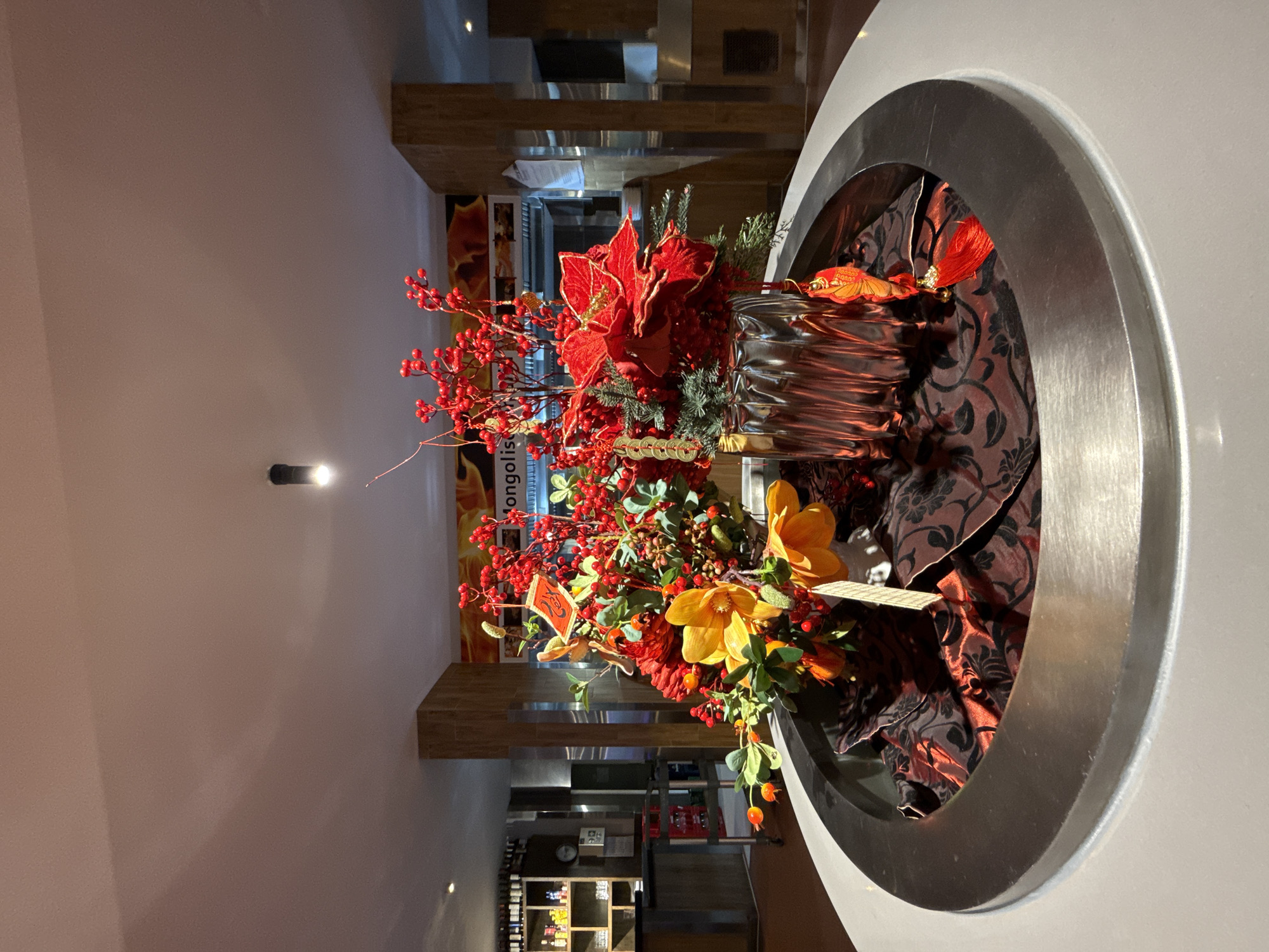 A still life of artificial floral arrangements is displayed on a circular, white surface within a metallic ring. The primary arrangement features red berries, large red poinsettias, and yellow flowers, all housed in a fluted silver vase. Chinese New Year decorations, including gold coins and red tassels, are incorporated into the arrangement. The background suggests an indoor commercial setting, likely a restaurant, with shelving units stocked with bottles on the left, and a kitchen area with stainless steel surfaces on the right. Text on a sign in the background reads "Mongolis...". The lighting appears to be artificial, with shadows cast on the ceiling. The overall impression is one of festive decoration within a dining establishment in Nordhausen, Germany.