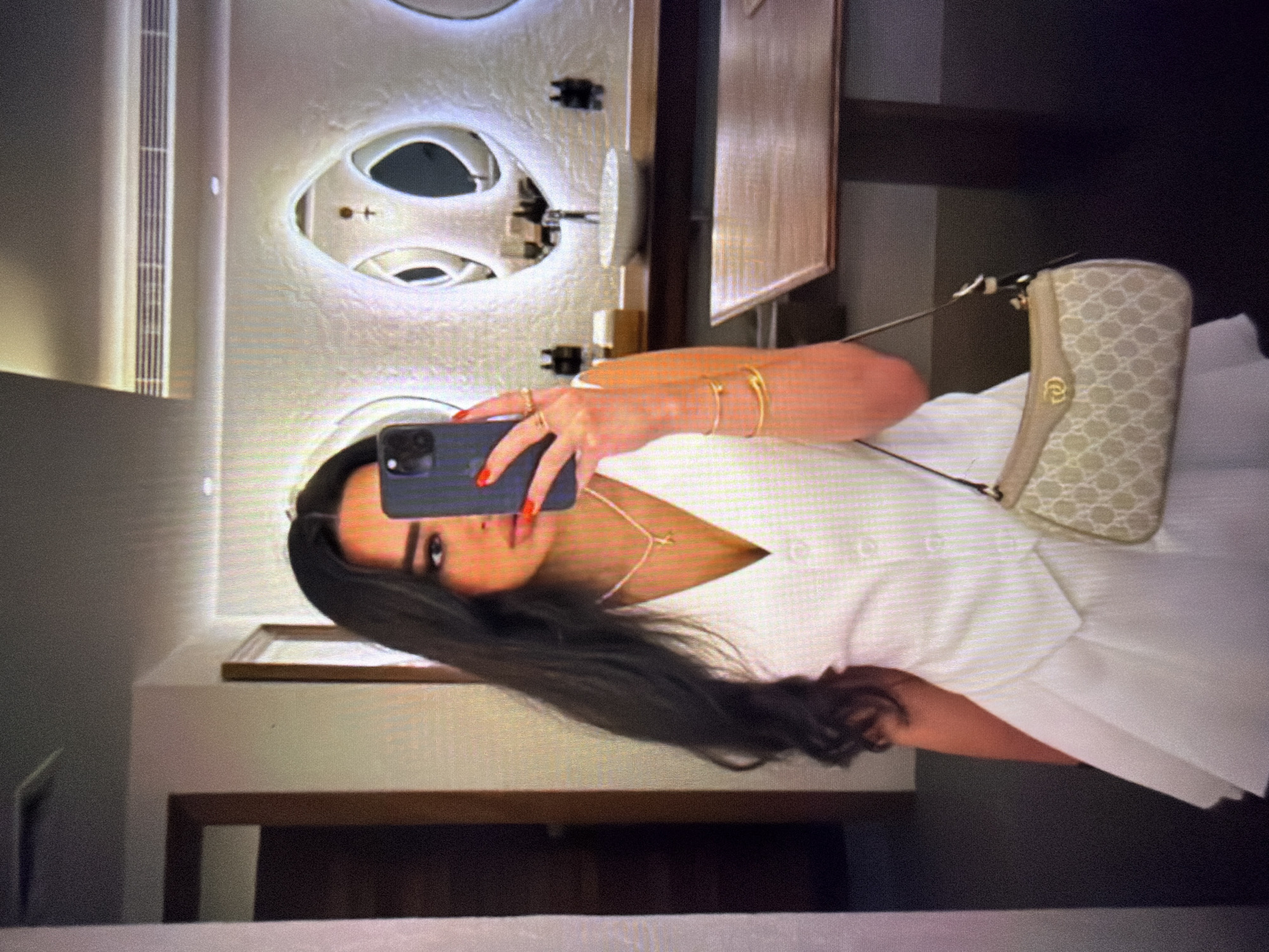 A woman with dark, wavy hair is taking a mirror selfie in what appears to be a modern bathroom. She is wearing a white vest over a white V-neck top and a white skirt. She has on a necklace with a small charm, rings on her fingers, and a bracelet. A beige and light brown patterned shoulder bag with a gold clasp hangs from her shoulder. The bathroom features a unique, sculpted white wall with irregularly shaped openings, one of which acts as a mirror reflecting the room and the woman. A sink with a faucet is visible on a dark countertop. A wooden table or counter is in the foreground. The background also shows a doorway to another room. The location is not specified in Beșalma, Moldova.