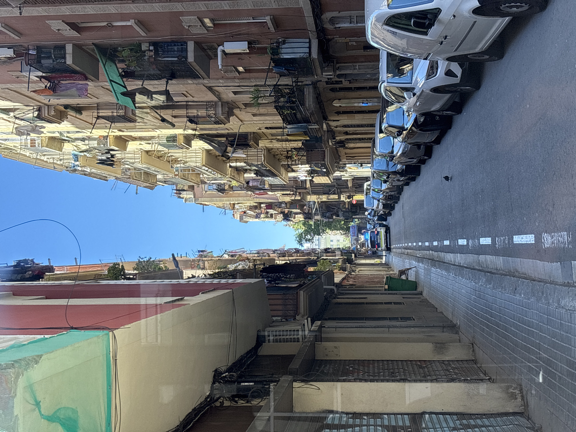 The image depicts a street scene in Barcelona, Spain, viewed from a slightly elevated position. The street is lined with multi-story buildings, many with balconies and clotheslines. Several cars are parked along the right side of the street, including white vans. The street appears to be narrow, with a clear blue sky visible overhead. A pigeon is on the street. Construction or renovation is visible in the foreground, with netting and building materials present. Wires are strung across some of the buildings.