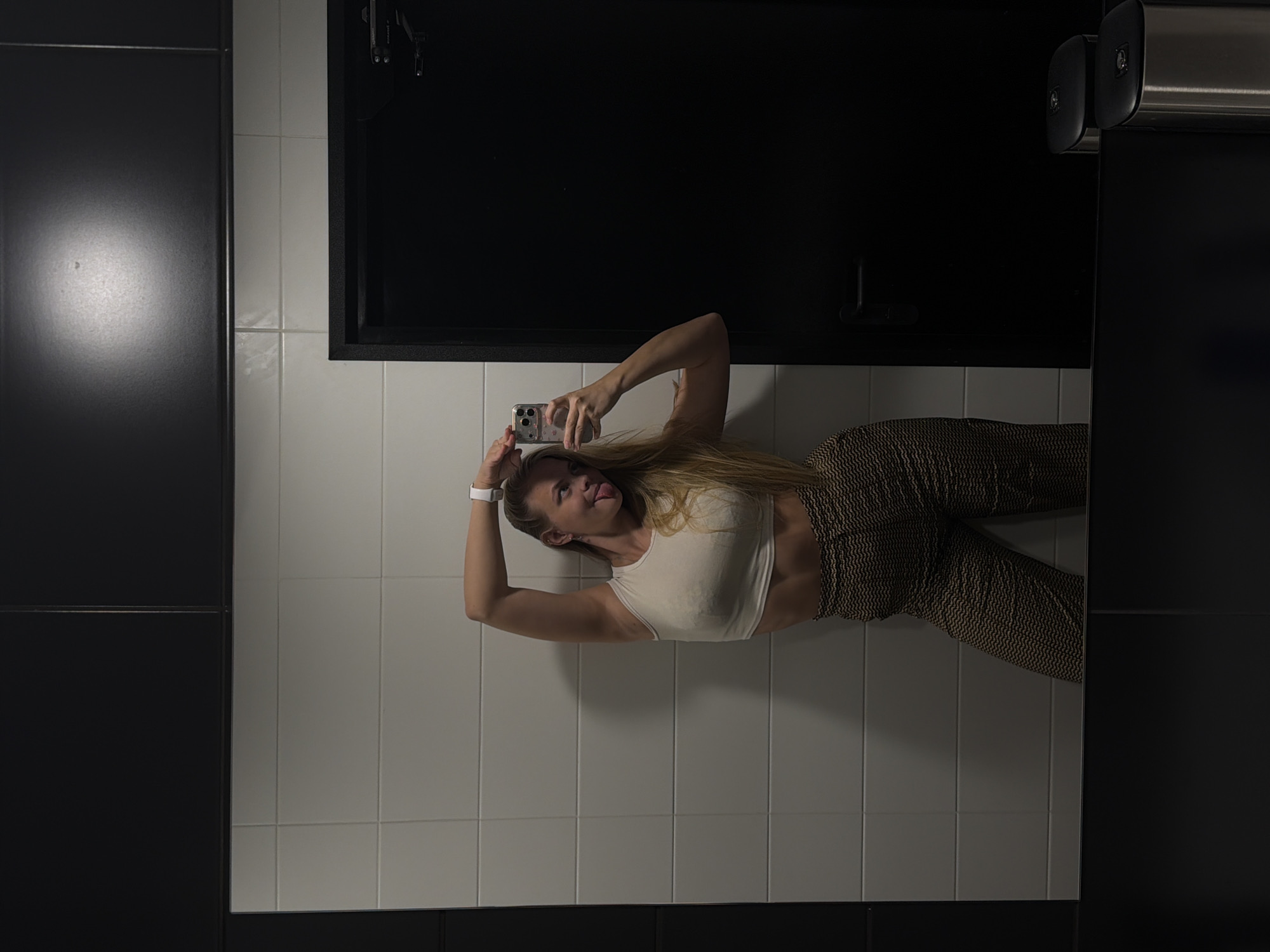 A woman is taking a selfie in a bathroom. She is wearing a white crop top and patterned pants. The bathroom has white tiled walls and a black door. The location is Izhevsk, Russia.