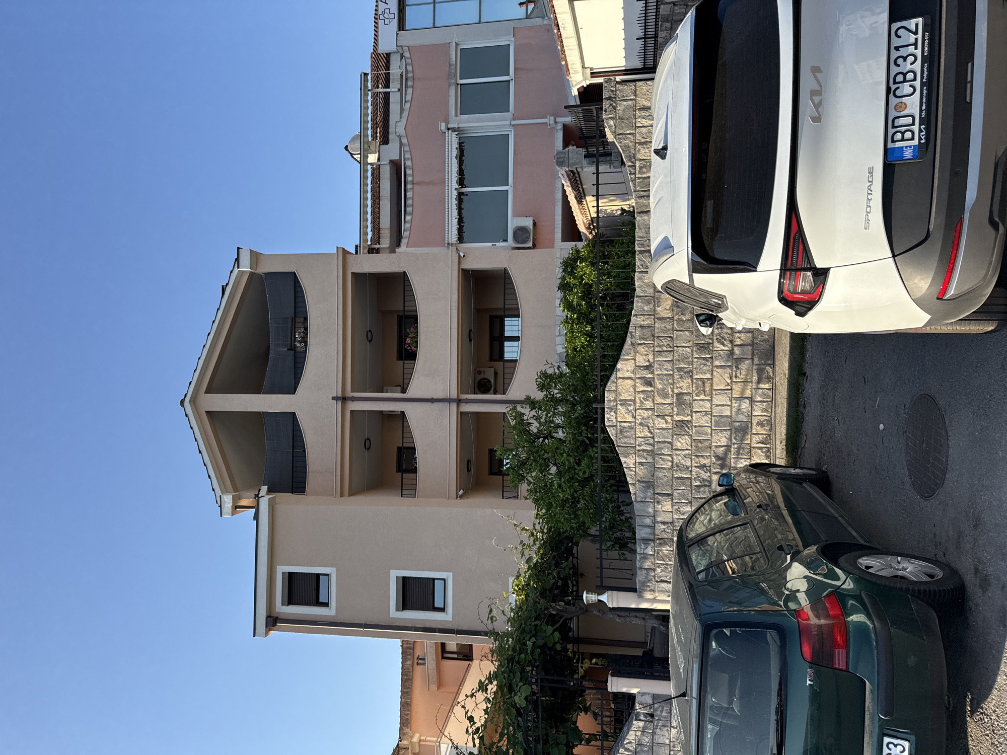 In Budva, Montenegro, a residential building with a light peach facade is visible under a clear blue sky. The building features multiple stories with balconies, some enclosed with dark mesh screens. On the ground level, a stone wall separates the property from a paved area. Two cars are parked in front of the wall: a dark green Volkswagen Golf TDI on the left and a white Kia Sportage on the right. The Kia Sportage has a license plate that reads "BD CB 312 MNE". Green foliage, including trees and vines, is growing behind the stone wall and the fence. The scene appears to be a quiet residential street, with the building likely housing apartments or a guesthouse.