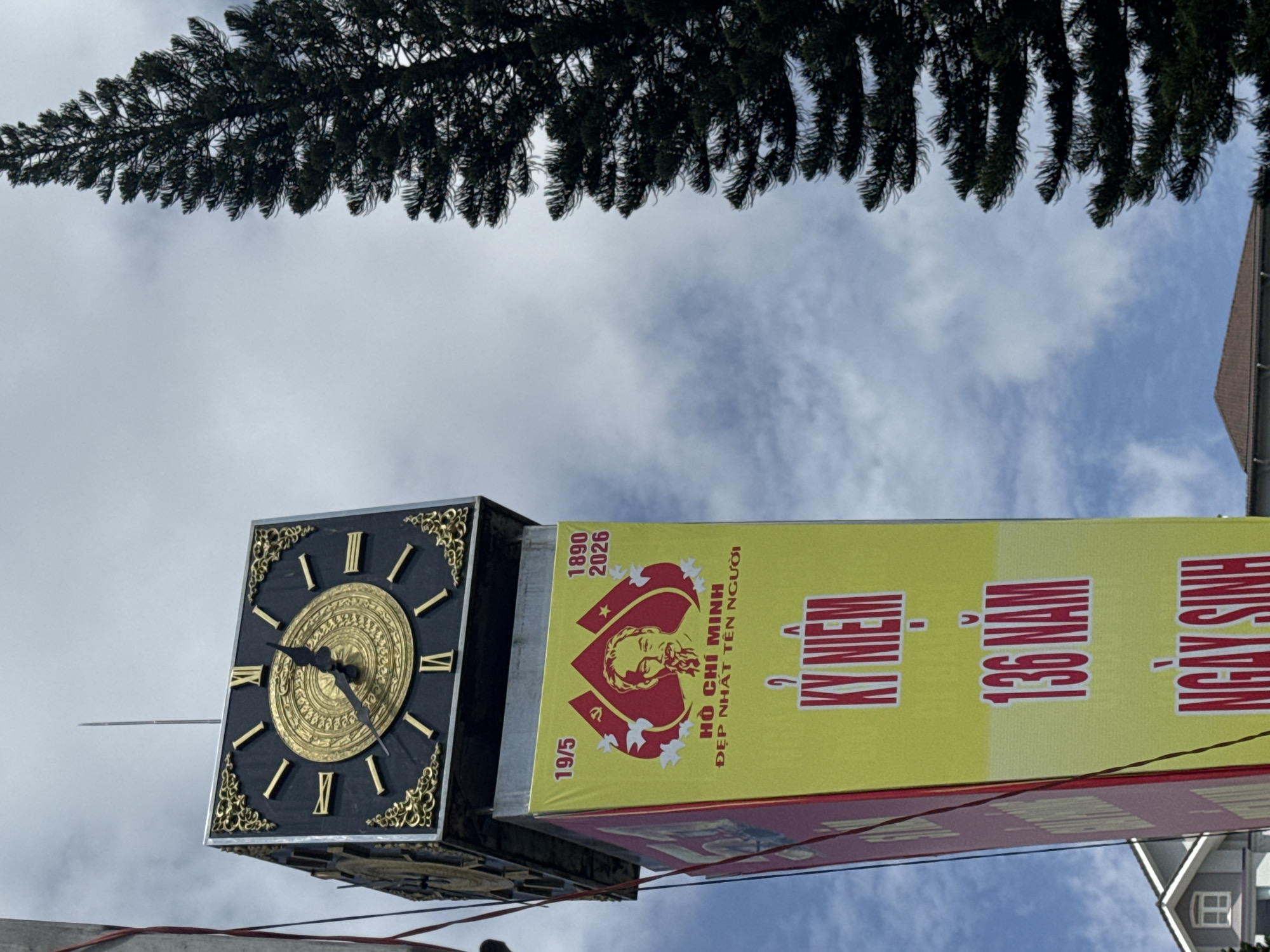 A tall clock tower in Da Lat, Vietnam, stands against a cloudy sky. A dark green pine tree is on the right side of the image. The clock tower has a black face with a gold, ornate clock and Roman numerals. Below the clock is a large yellow banner with red text and an image of Ho Chi Minh. The banner reads "19/5 1890-2026 HO CHI MINH DEP NHAT TEN NGUOI KY NIEM 136 NAM NGAY SINH". To the left of the banner is a red banner with some text that is not fully visible. In the background, on the left side of the image, are parts of buildings with tiled roofs.