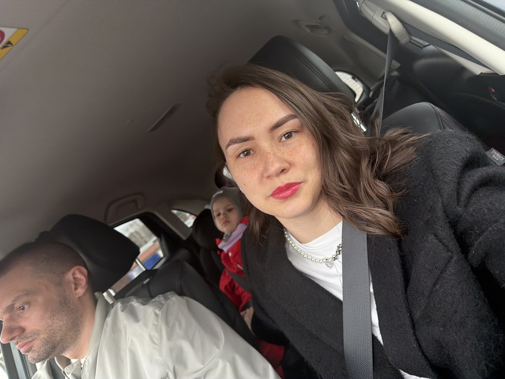 A medium shot taken inside a car, likely in motion, features three individuals. A fair-skinned woman with brown hair and visible freckles is in the foreground, wearing a black cardigan over a white t-shirt adorned with a pearl necklace, and her lips are painted red. A seatbelt is secured across her chest. Behind her and to the left sits a child in a red jacket and grey knit hat, looking towards the camera. To the woman's left and further in the background, a man with fair skin and a shaved head is visible, wearing a light grey jacket, and he is looking towards the car window on the left. The interior of the car is visible, including the seats, headrests, and parts of the dashboard and windows. Outside the windows, blurry cityscapes are visible, suggesting the car is moving through an urban environment, specifically Chaikovsky, Russia.