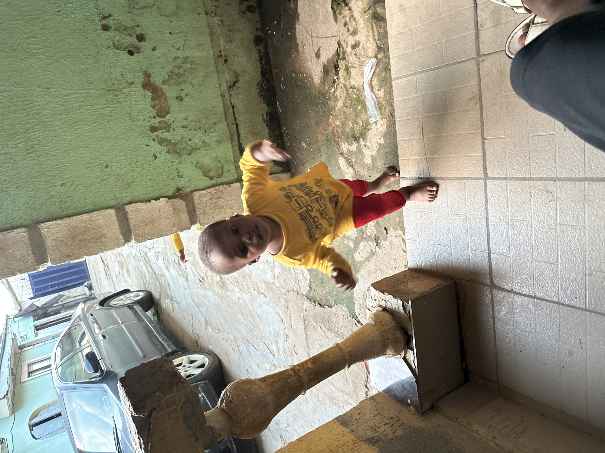 A young child in a yellow shirt and red leggings stands on a tiled entryway in Narayi, Nigeria. The child is looking towards the camera with their left hand outstretched. A dark sedan is parked in the background on a dirt and concrete street. The building to the right has a textured green wall.