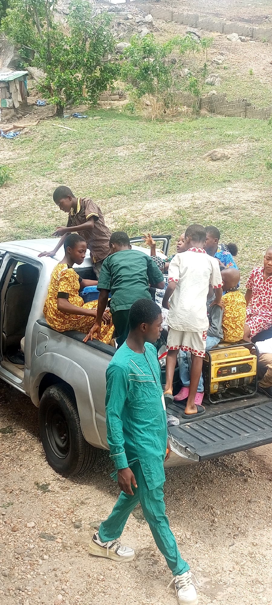 The scene depicts a group of people in and around a silver pickup truck. The truck's bed is occupied by several children and a portable generator. One person in a green suit is standing next to the truck and another person is leaning over the side. The location appears to be an outdoor area with sparse vegetation and rocky terrain. Given the prompt, it is plausible that this scene is in Lokoja, Nigeria.